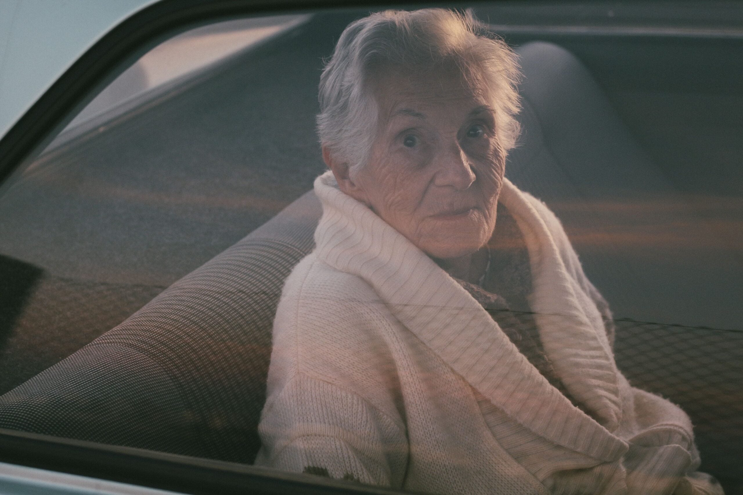 An elderly woman with short gray hair and a white sweater sits in the backseat of a car, looking out the window with a calm expression.