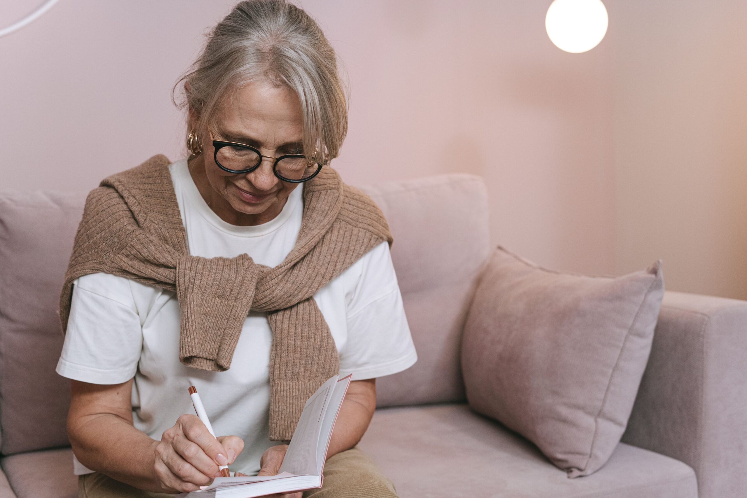 An older woman with glasses sits on a sofa, writing in a small notebook.
