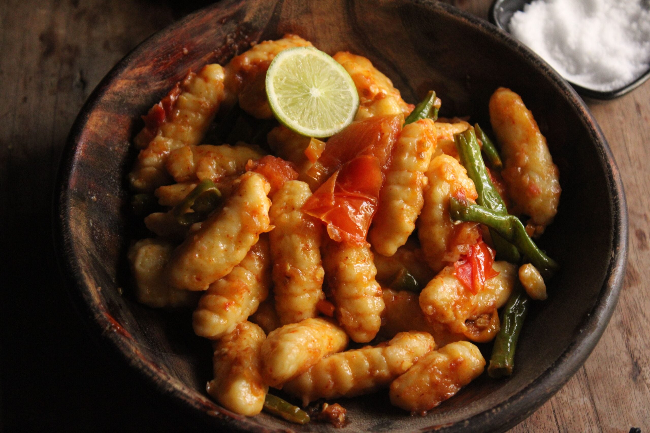 Gnocchi tossed in a red sauce with green beans and a lime slice in a wooden bowl.