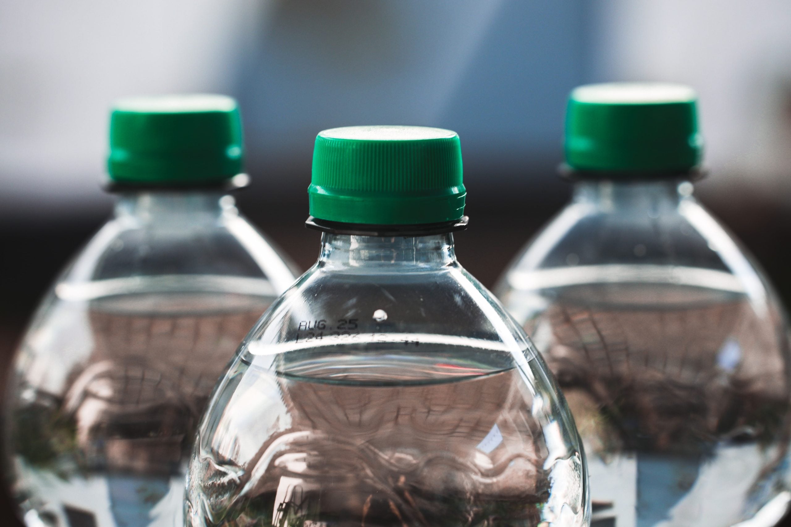 Close-up of three clear plastic water bottles with green caps, positioned side by side. The background is blurred, focusing attention on the tops of the bottles in the foreground.