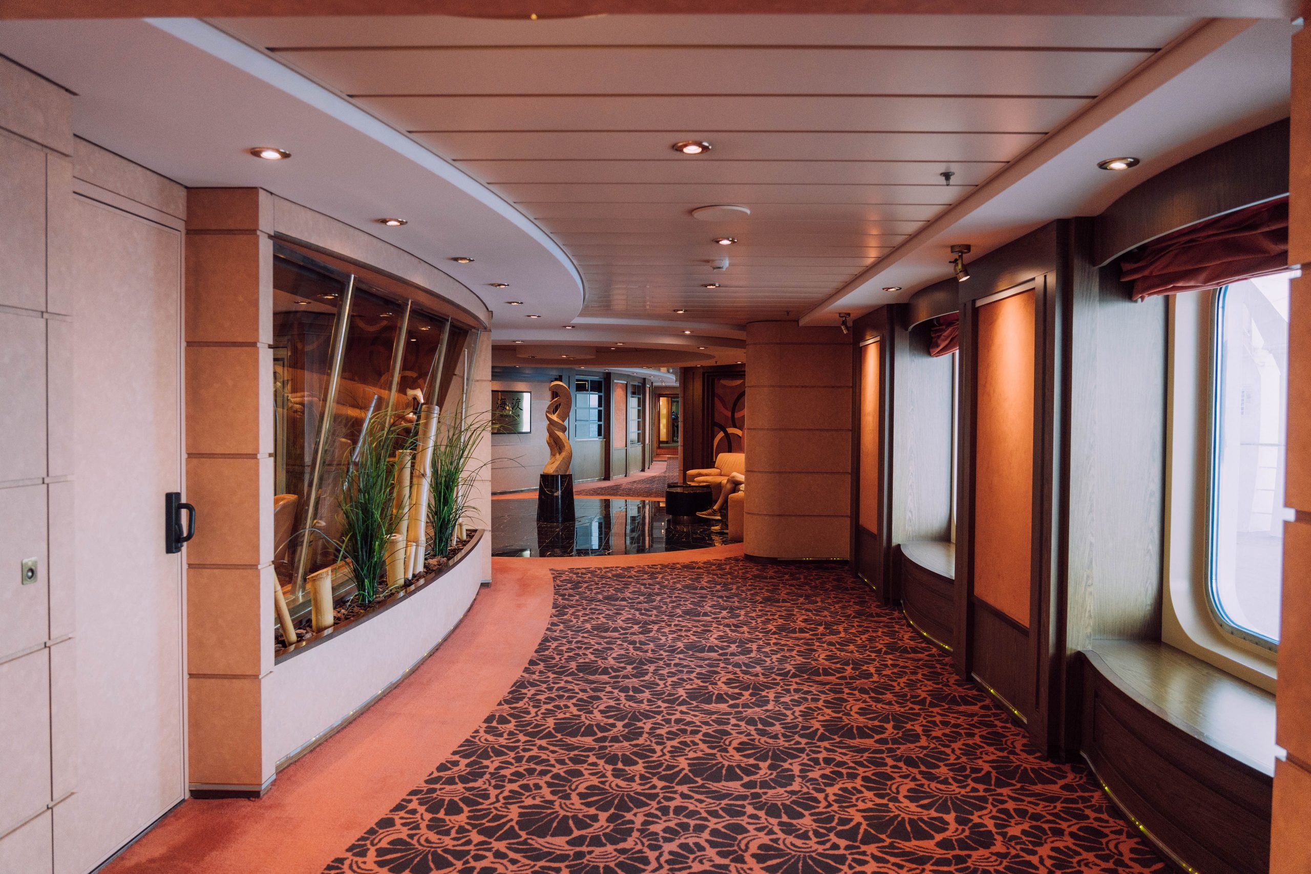 A curved hallway with patterned carpet, large windows on the right, and decorative plants on the left, leading to a sitting area with sculptures and chairs in the background.