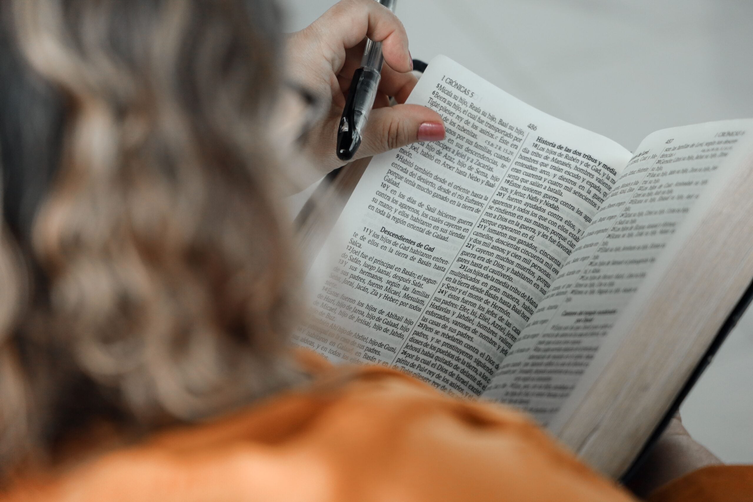 A person holding an open Bible and a black pen, reading the text on the page.