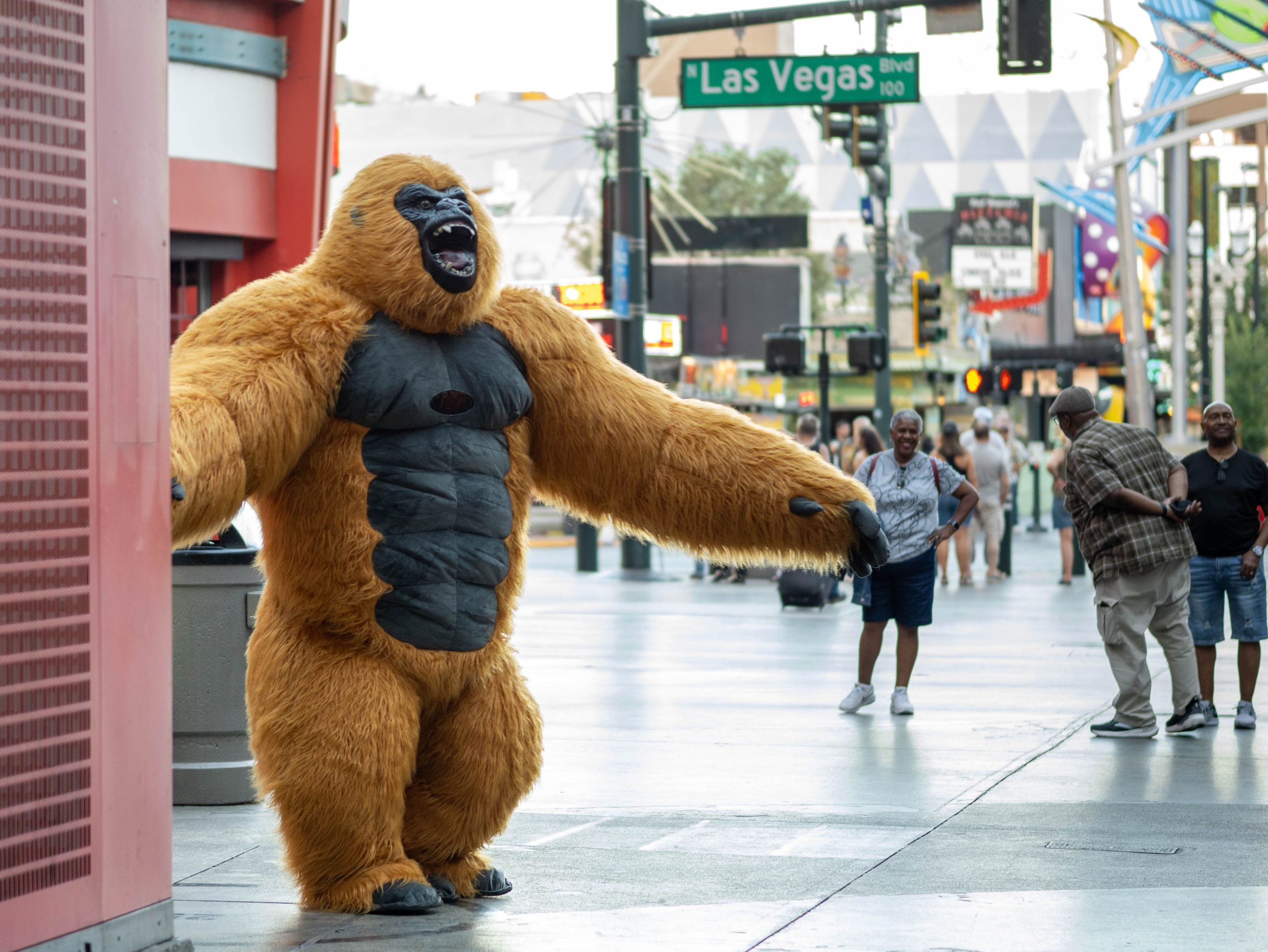 Person in an orange and black gorilla costume poses with arms wide open on a busy street in Las Vegas, with onlookers and city signs in the background.