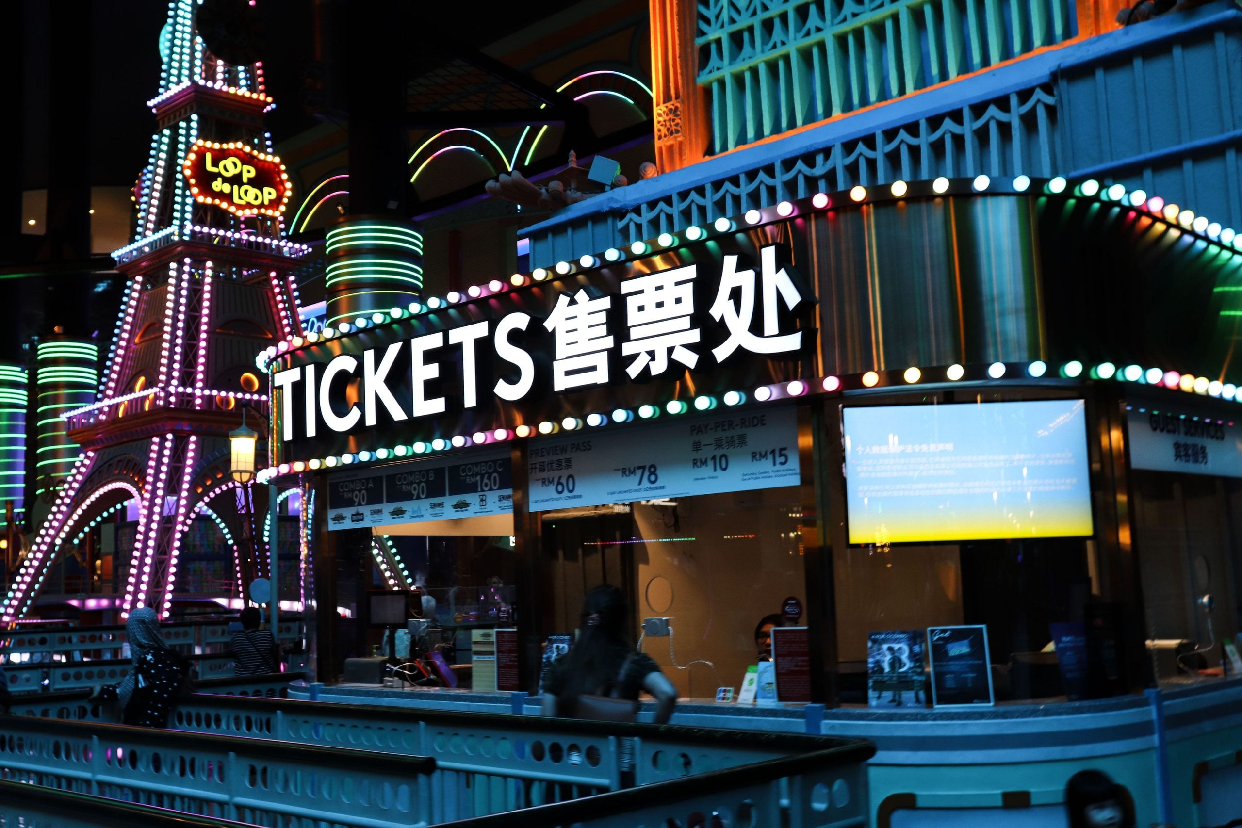 A brightly lit ticket booth with “TICKETS” and Chinese characters above the window, surrounded by colorful neon lights and a replica Eiffel Tower in the background.