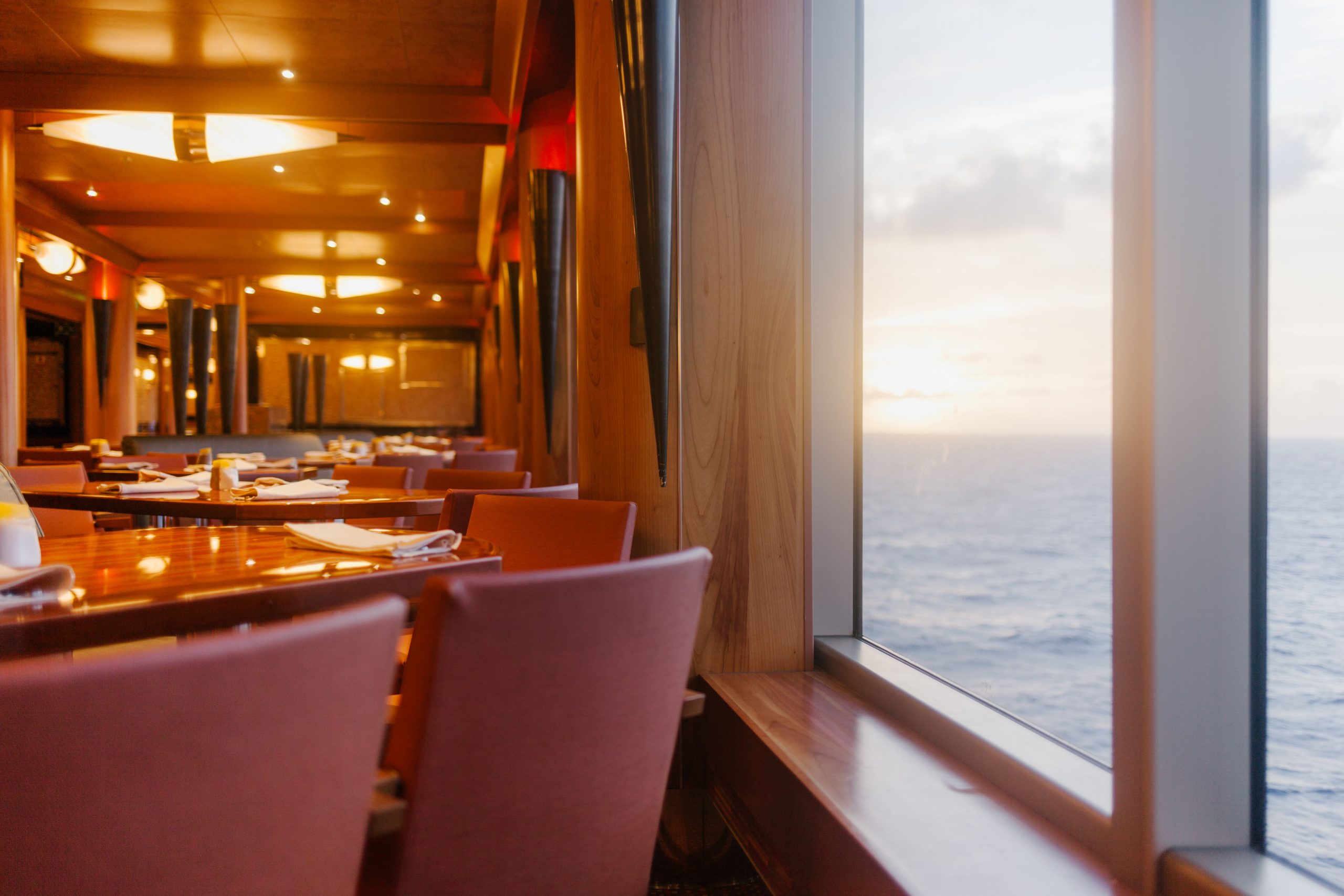 Elegant restaurant with pink chairs and set tables beside large windows overlooking the ocean at sunset, with warm lighting creating a cozy atmosphere.