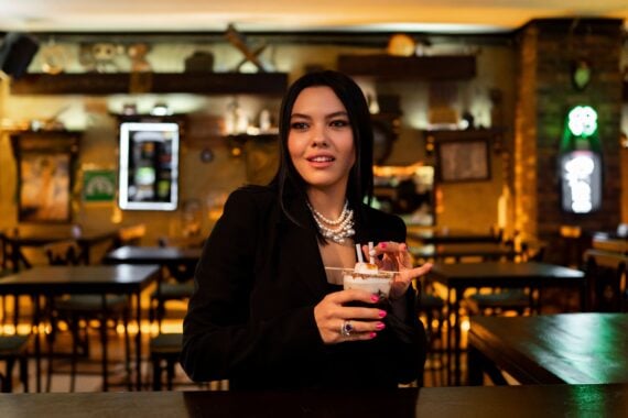 A woman wearing a black blazer and pearl necklace holds a cocktail with a toasted marshmallow garnish in a dimly lit bar.