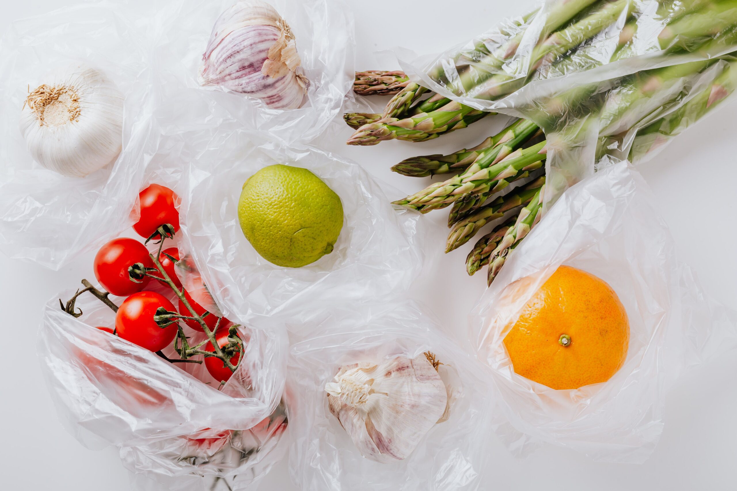 Various fresh vegetables and fruits, including tomatoes, garlic, lime, orange, and asparagus, are arranged on a white surface, each wrapped in clear plastic bags.