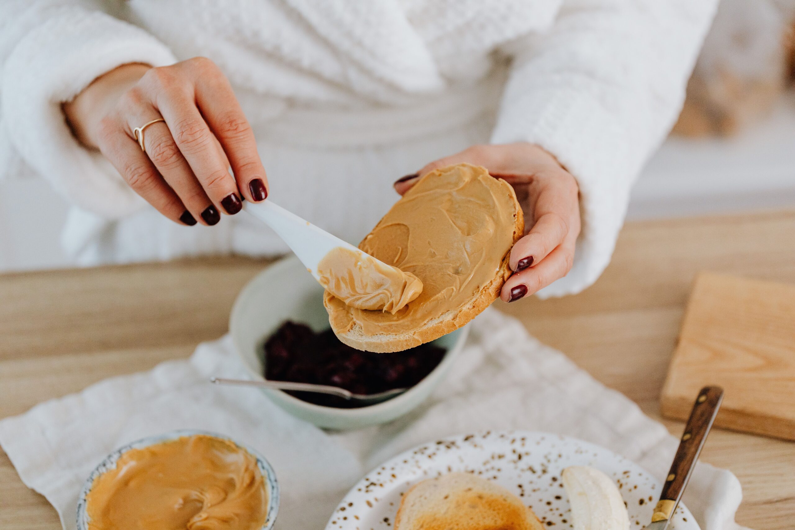 A person in a white robe spreads peanut butter on a piece of toast with a white knife. There are bowls of peanut butter and jam, a plate with toast, and a banana on the table.