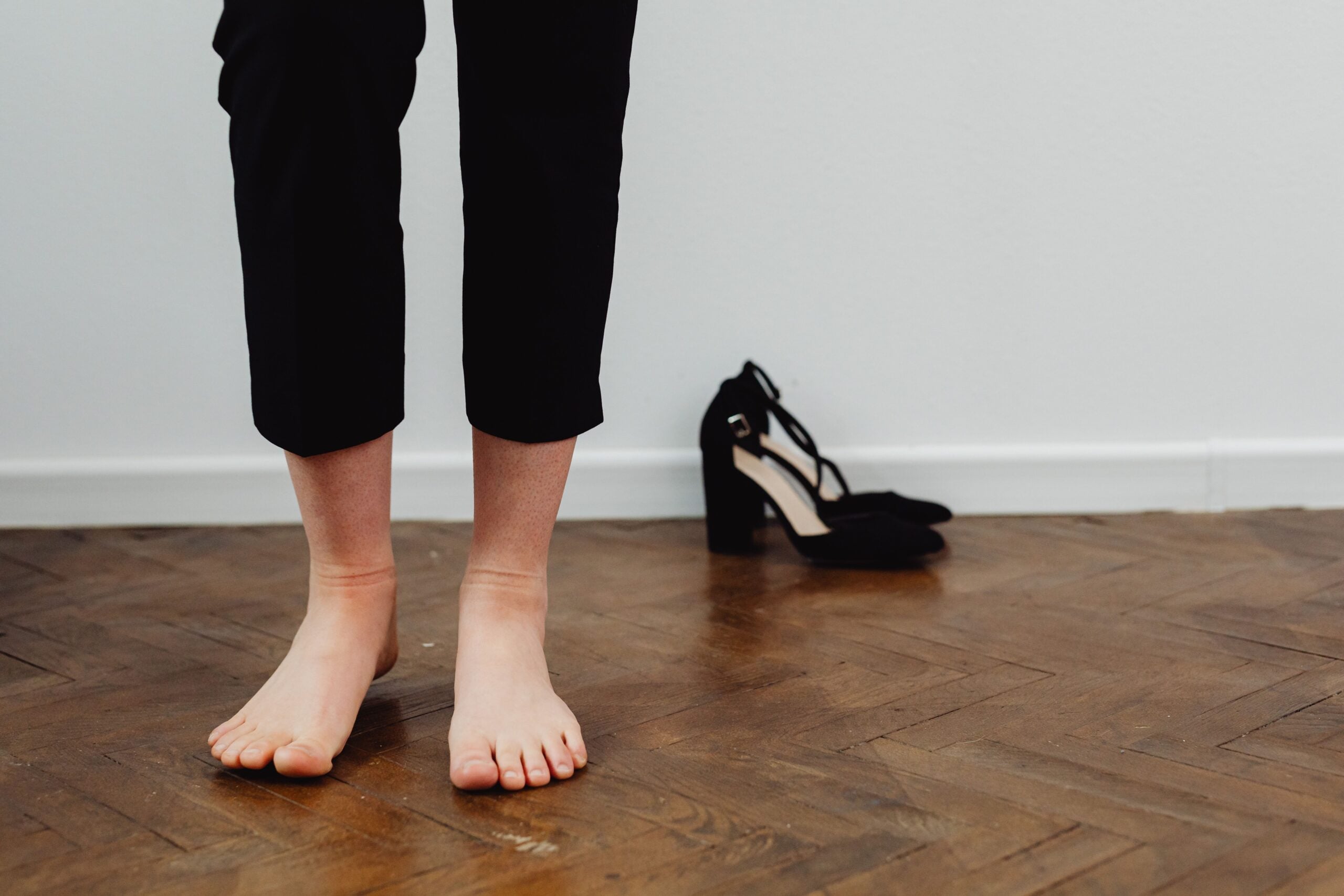 A person standing barefoot on a wooden floor in black trousers, with a pair of black high-heeled shoes resting on the floor behind them.