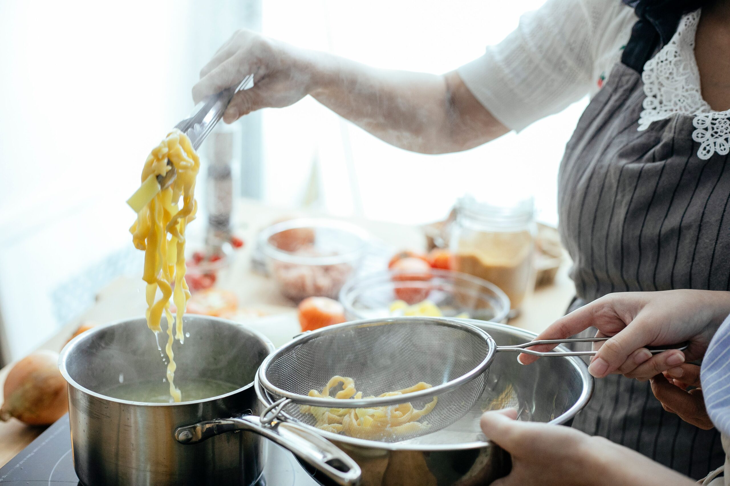 Two people cook together, using tongs to lift boiled pasta from a pot and transfer it into a metal strainer over another pot. Ingredients and utensils are visible on the kitchen counter nearby.