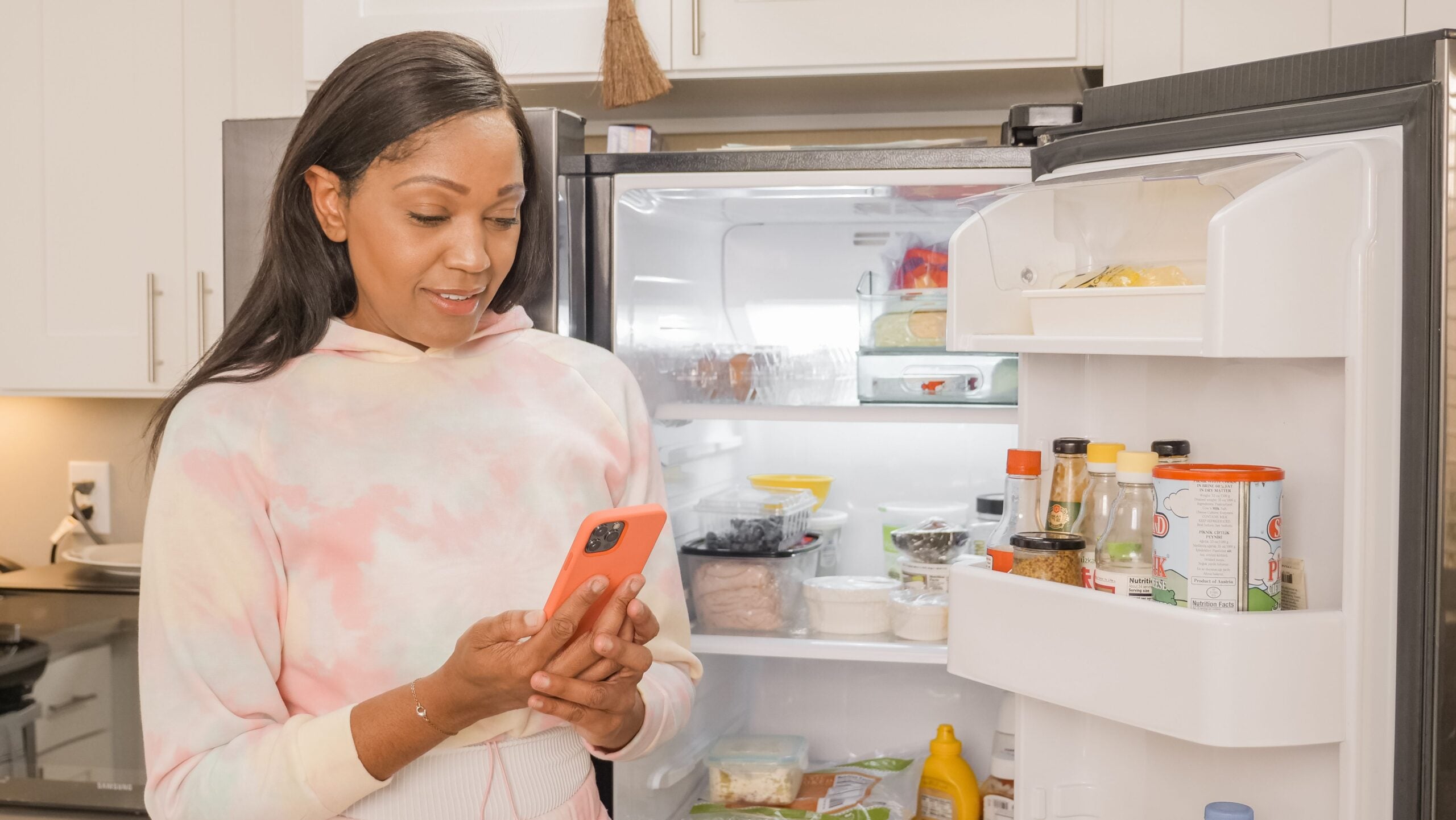 A woman in a tie-dye sweatshirt looks at her smartphone while standing in front of an open refrigerator.