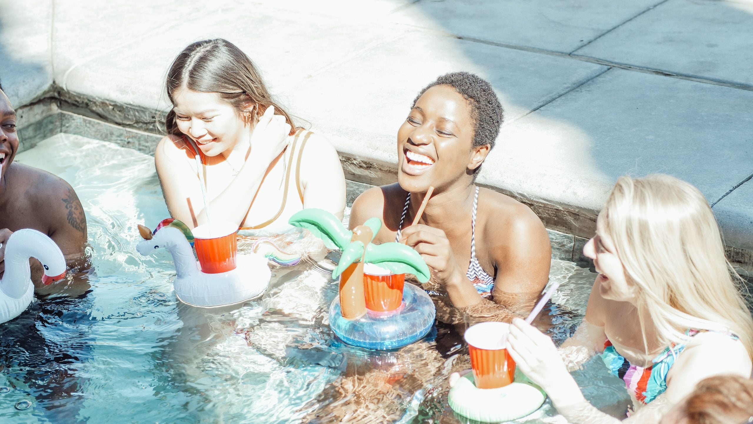 Four friends sit in a pool, laughing and holding inflatable drink holders with red cups. Sunlight shines on them, creating a bright, cheerful atmosphere.