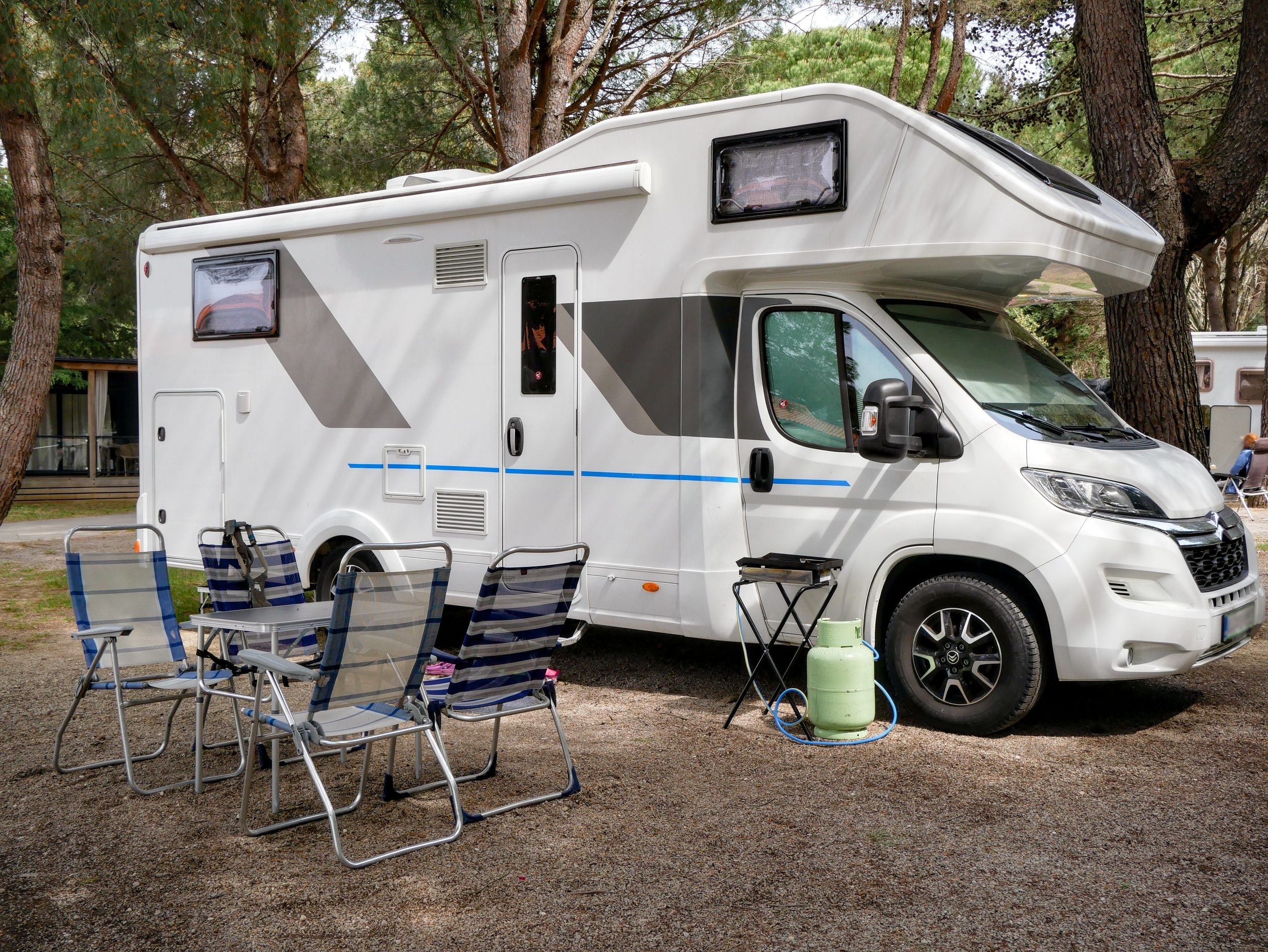A white camper van is parked among trees. In front of it, there are four folding chairs and a table. A green gas canister and a small stool are beside the camper van. The setting appears to be a campsite.