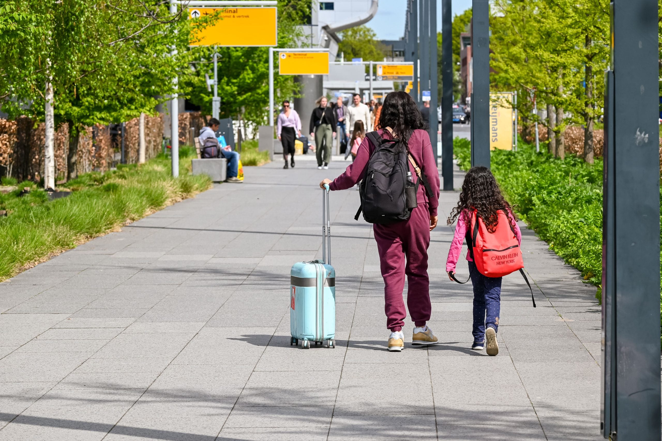 A woman pulling a light blue suitcase and a young girl with a red backpack walk away from the camera along a paved outdoor walkway lined with trees.