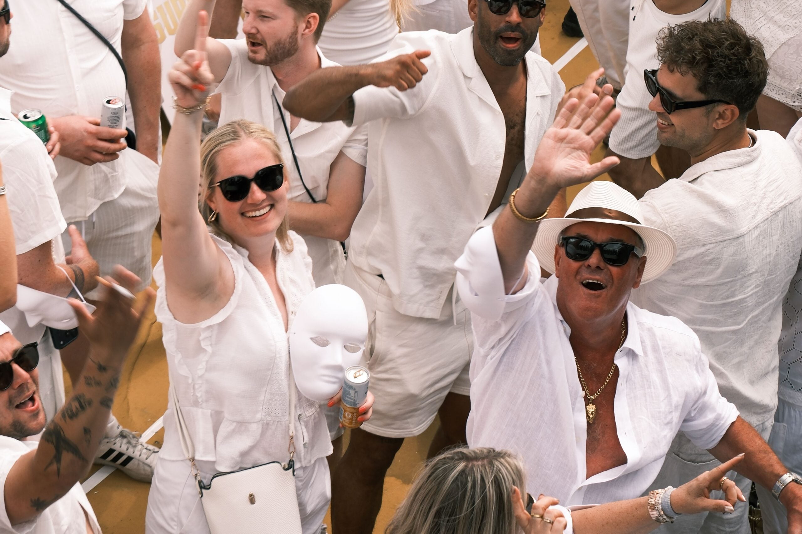 A crowd dressed in white clothing celebrating at an outdoor cruise entertainment event, with several individuals waving to the camera.