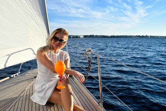 A woman in a white dress sits on a sailboat deck, holding an orange drink, smiling, and wearing sunglasses. Blue water and a distant shoreline are visible under a clear sky.