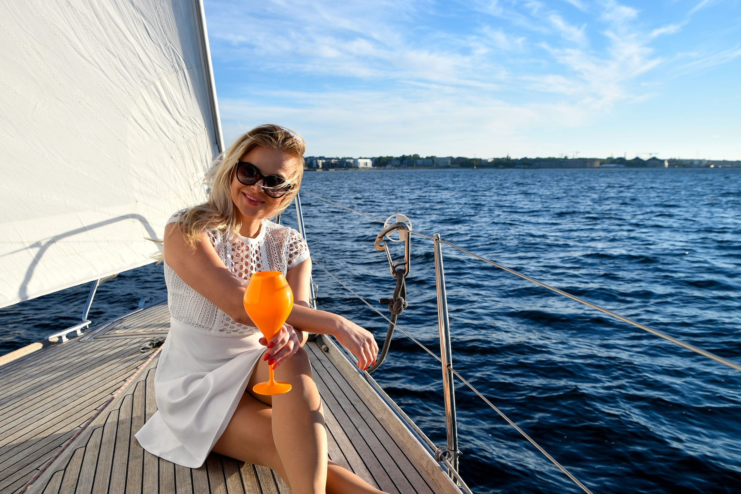A woman in a white dress sits on a sailboat deck, holding an orange drink, smiling, and wearing sunglasses. Blue water and a distant shoreline are visible under a clear sky.