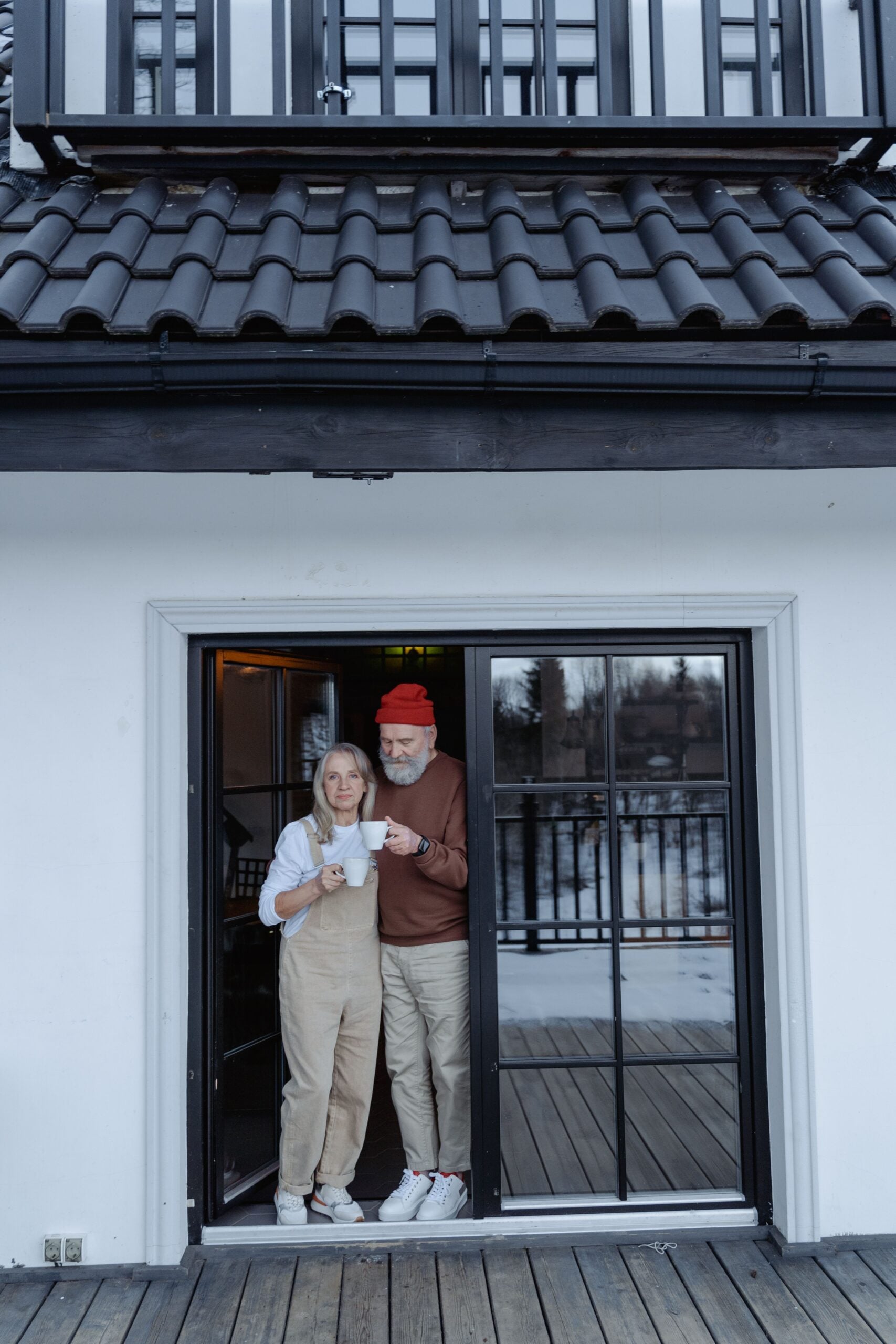 An older couple stands in a doorway, holding mugs and enjoying a hot drink together. The woman wears overalls; the man wears a red beanie. They are on a wooden deck outside a white house with a black roof.