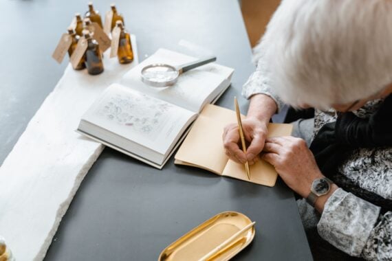 A person writes in a notebook at a table with an open book, a magnifying glass, and several small amber glass bottles.