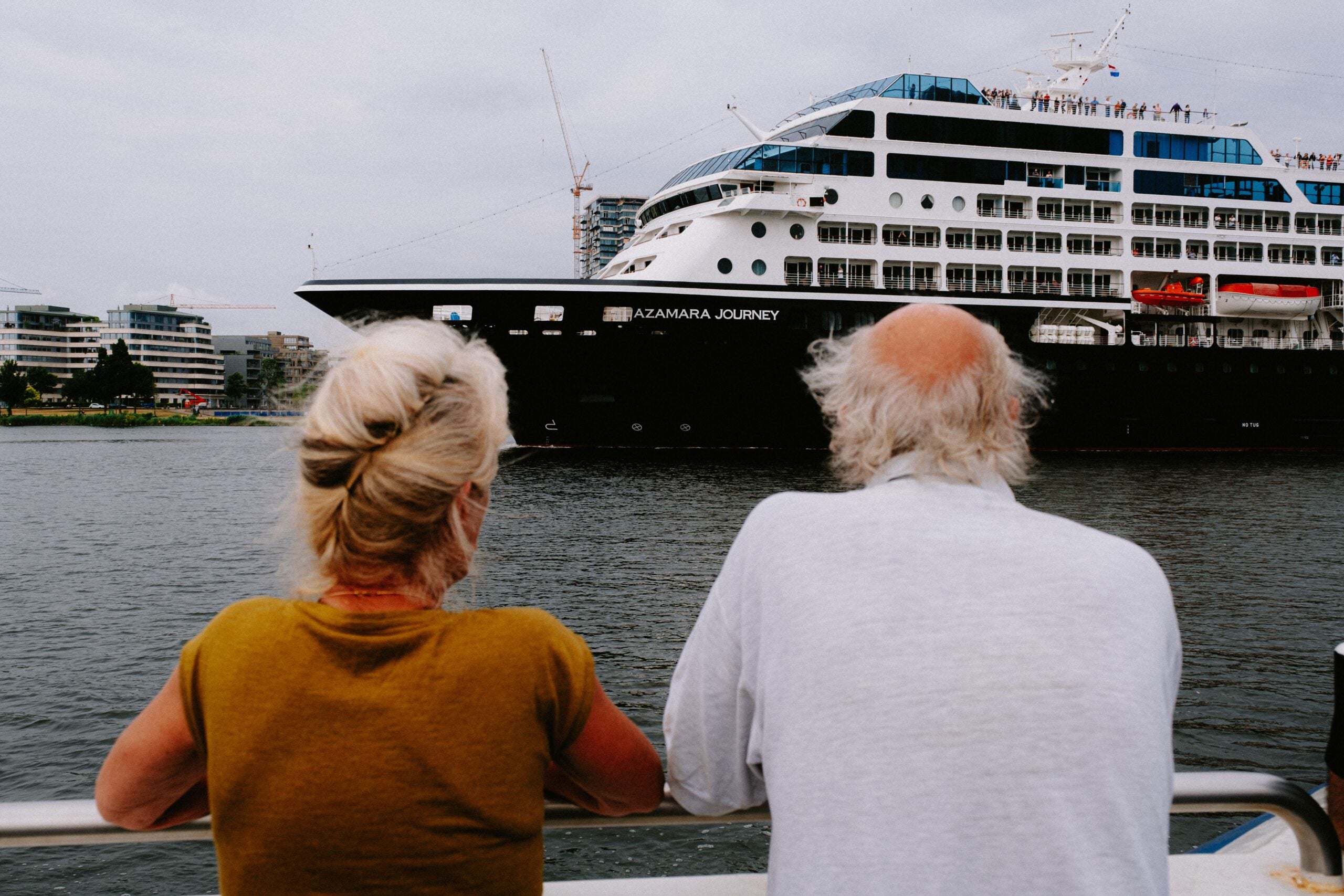 An older woman and man with gray hair stand by a railing, looking at a large cruise ship named “Azamara Journey” passing on the water near city buildings.
