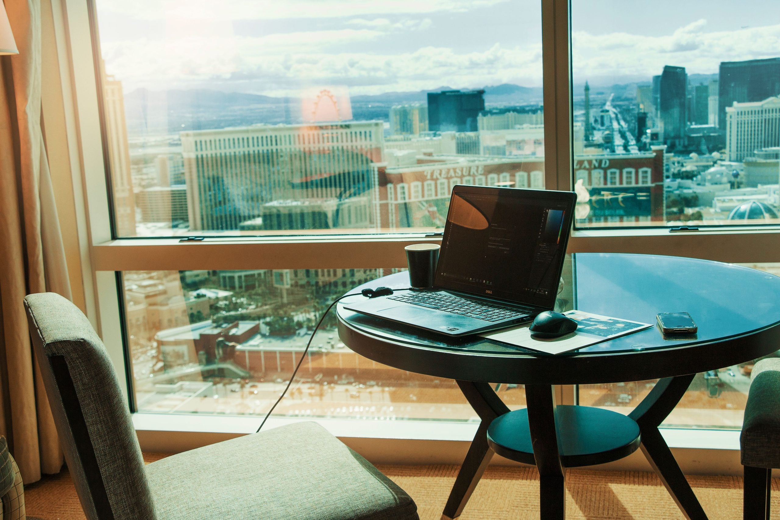 A laptop, mouse, cup, and phone on a round table by a window with a city skyline view. Two chairs face the table, and sunlight streams through the window, illuminating the workspace.