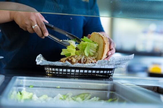A person in a blue shirt uses tongs to place lettuce onto a sandwich in a basket lined with printed paper.