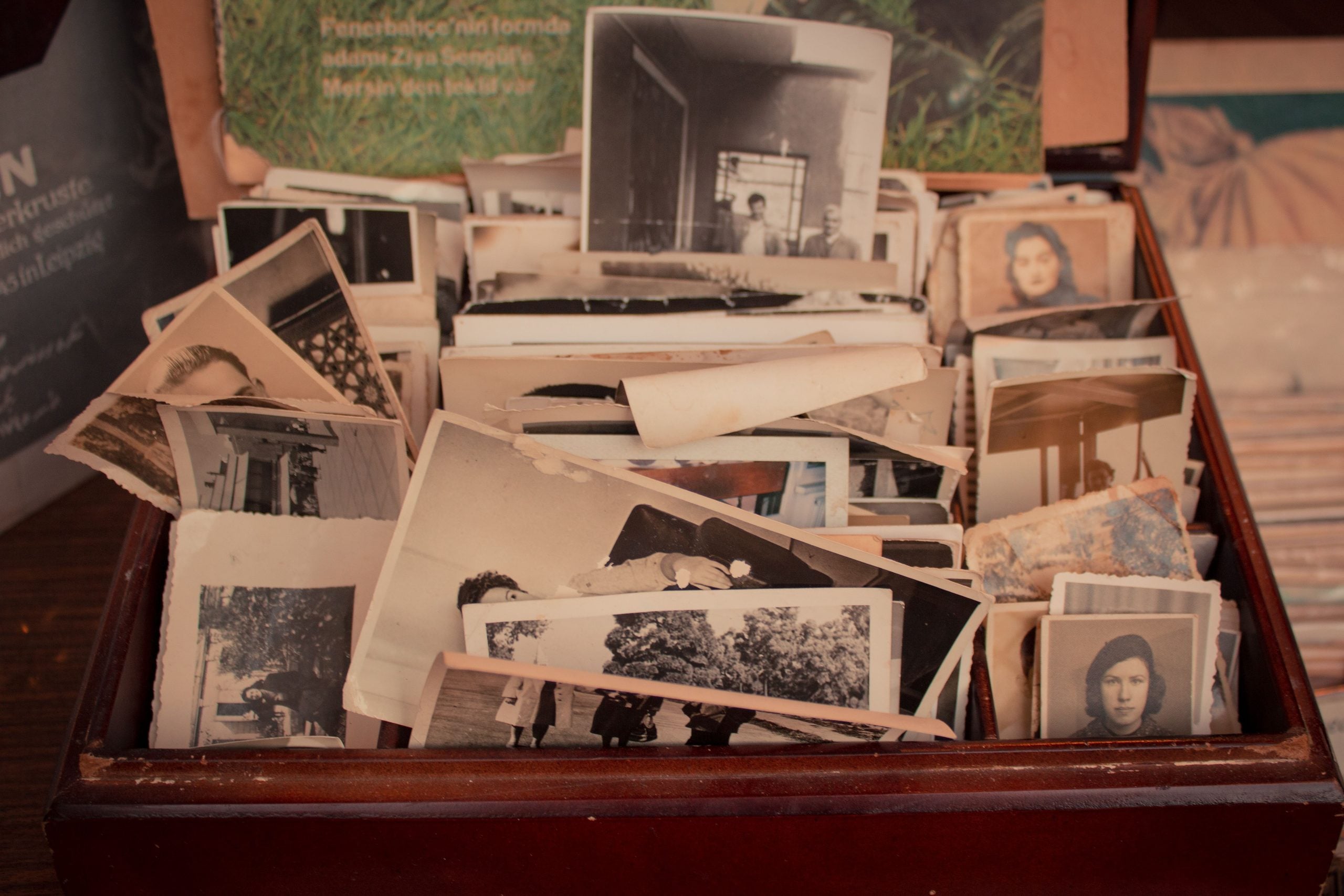 An open box filled with old black-and-white photographs, some slightly curled, showing various people and scenes. The photos are stacked and overlapping, evoking a sense of nostalgia and history.