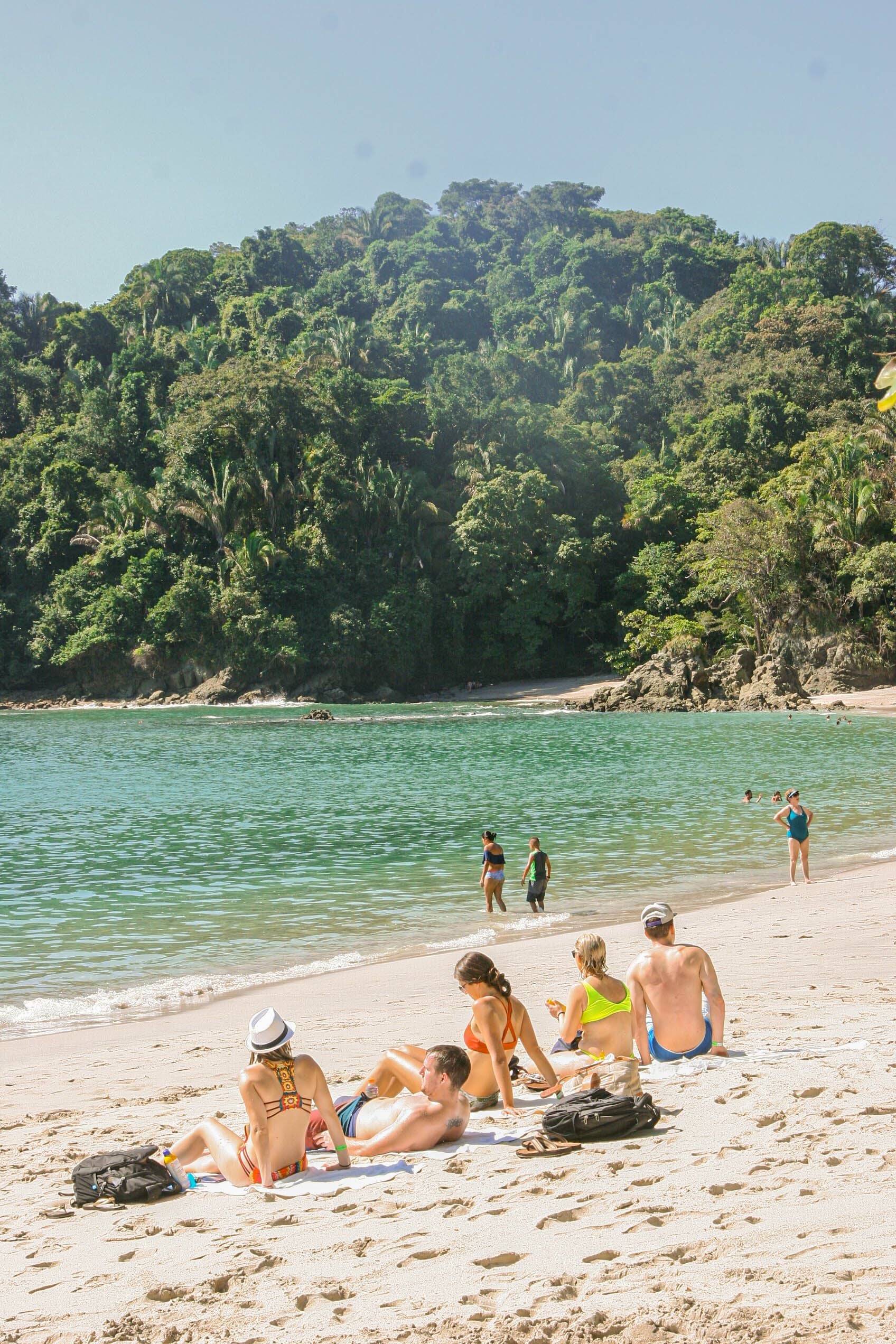 People relax on a sandy beach near clear green water and lush green hills. Some are lying on towels while others walk by the shore under a clear sky.