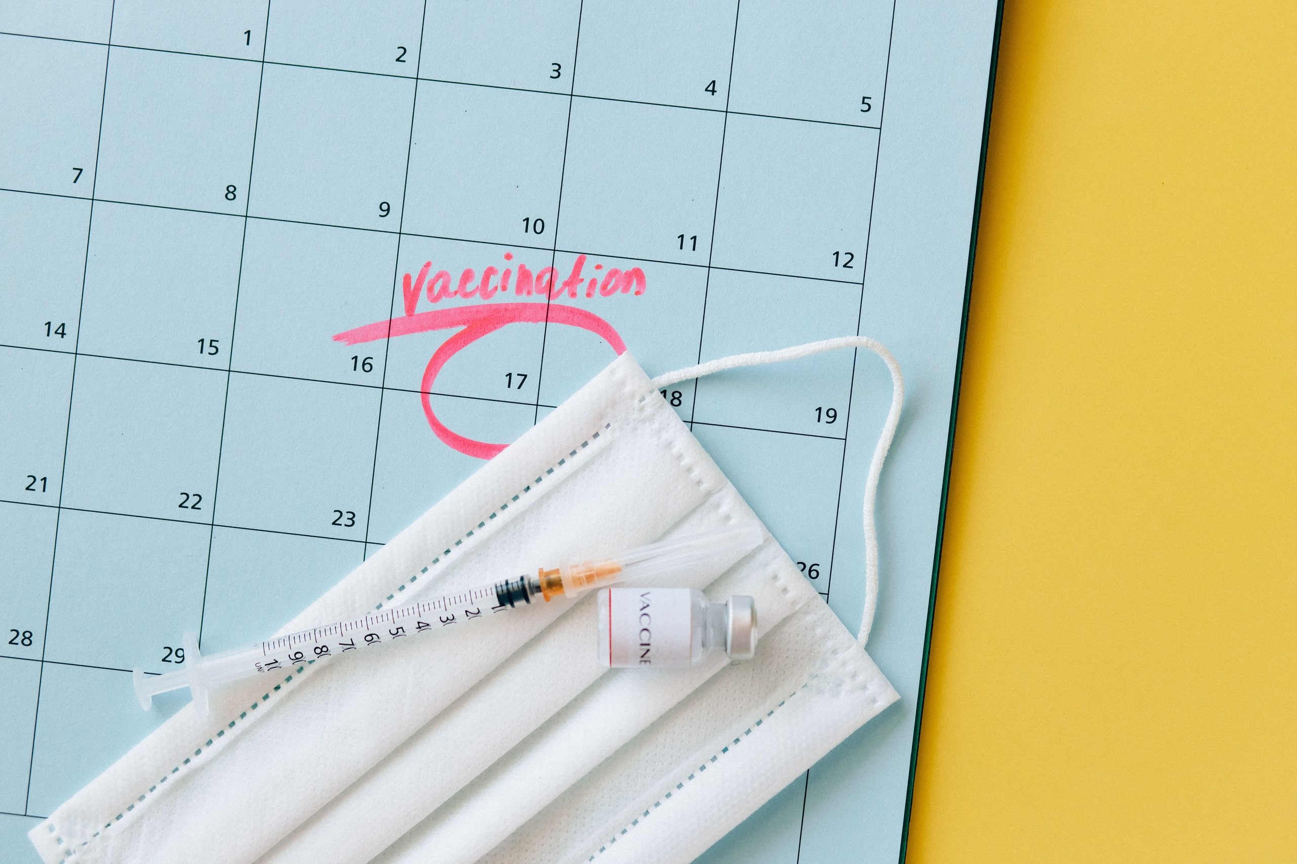 A face mask, syringe, and vaccine vial resting on a calendar with the 17th circled in red and labeled “Vaccination.”