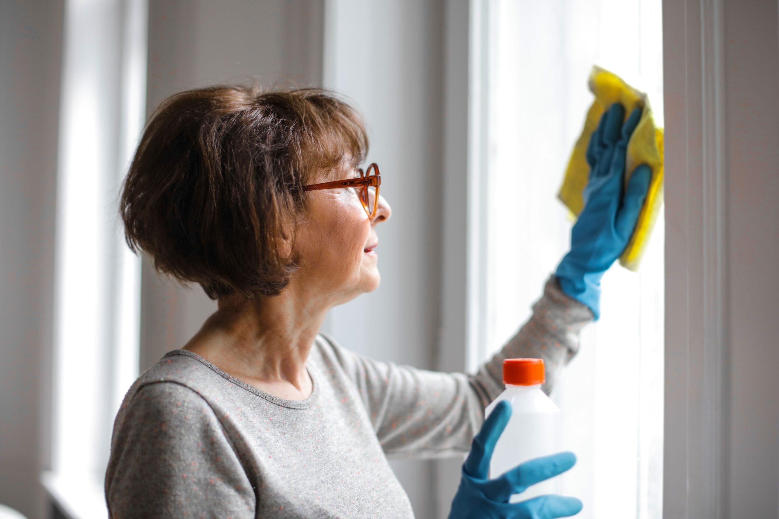 An older woman wearing glasses and blue gloves cleans a window with a yellow cloth, holding a bottle of cleaning solution in her other hand.