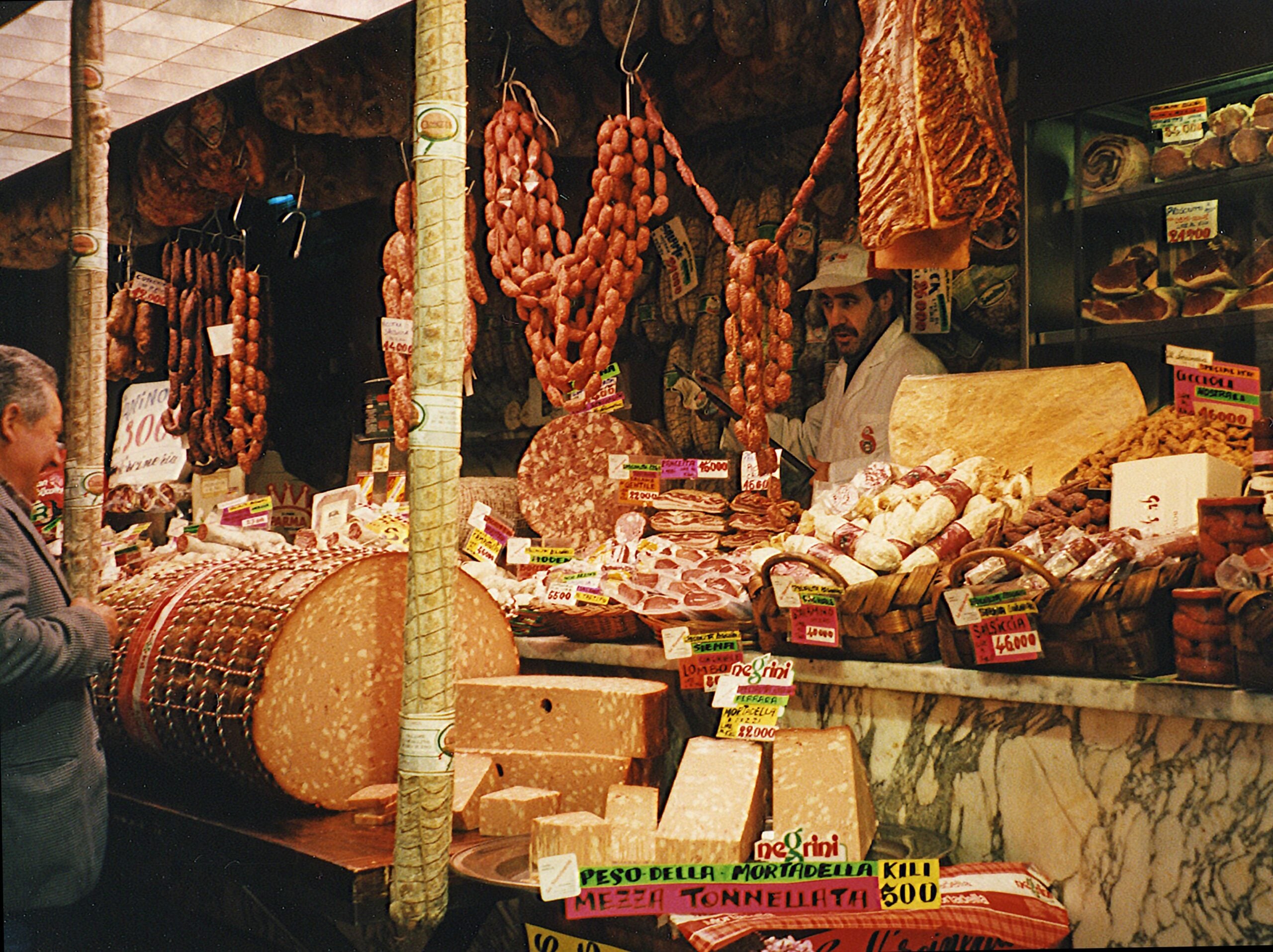A shop attendant behind a counter filled, in one of the specialty stores, with large wheels of mortadella, various cheeses, and assorted hanging sausages, with price tags displayed throughout.