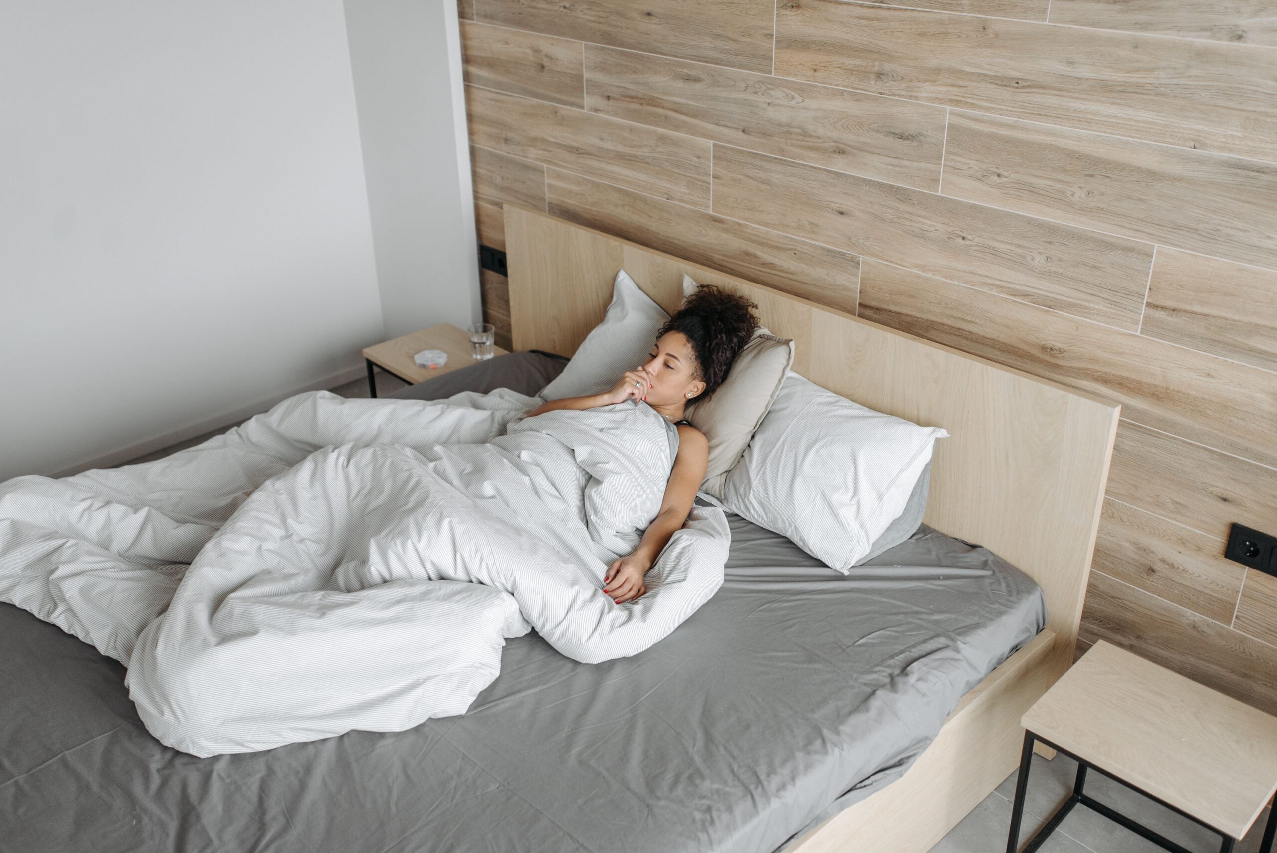 A woman with curly hair lies in bed under a white duvet, coughing into her hand.