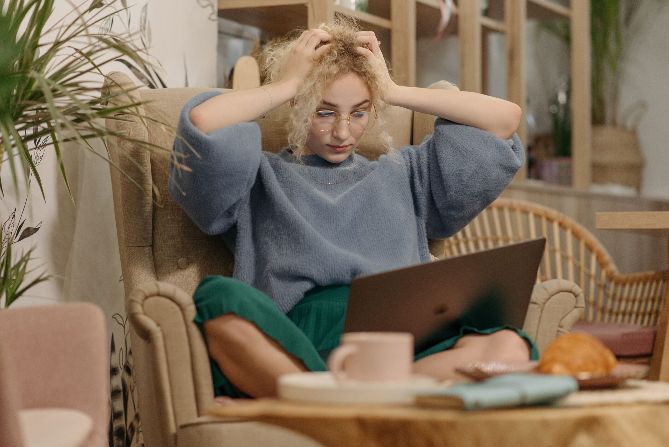 A person with curly blonde hair and glasses sits in an armchair with their hands on their head, looking stressed while working on a laptop.