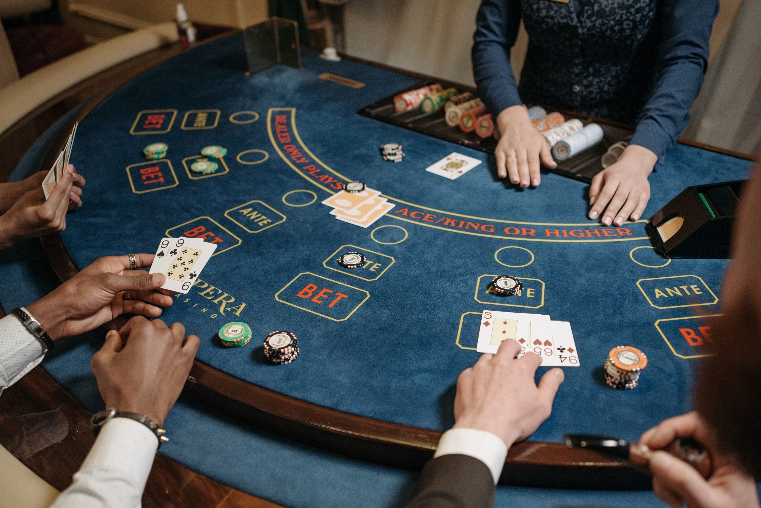 Three people play cards at a casino table with poker chips, cards, and a dealer in a blue vest. The table has labeled sections for bets, “ANTE,” and “ACE/KING OR HIGHER.”