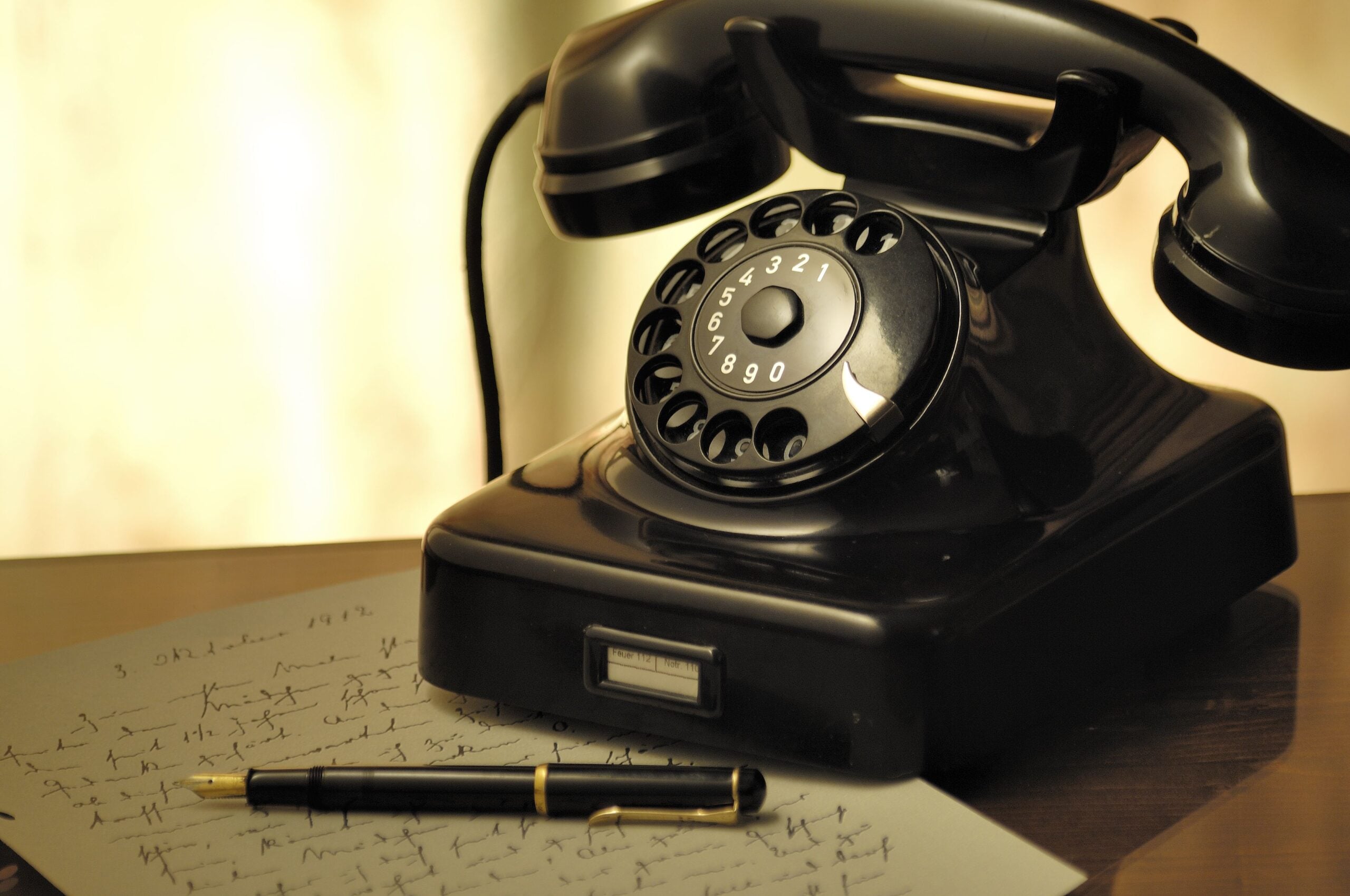 A black rotary telephone sits on a desk next to a fountain pen resting on a handwritten letter.