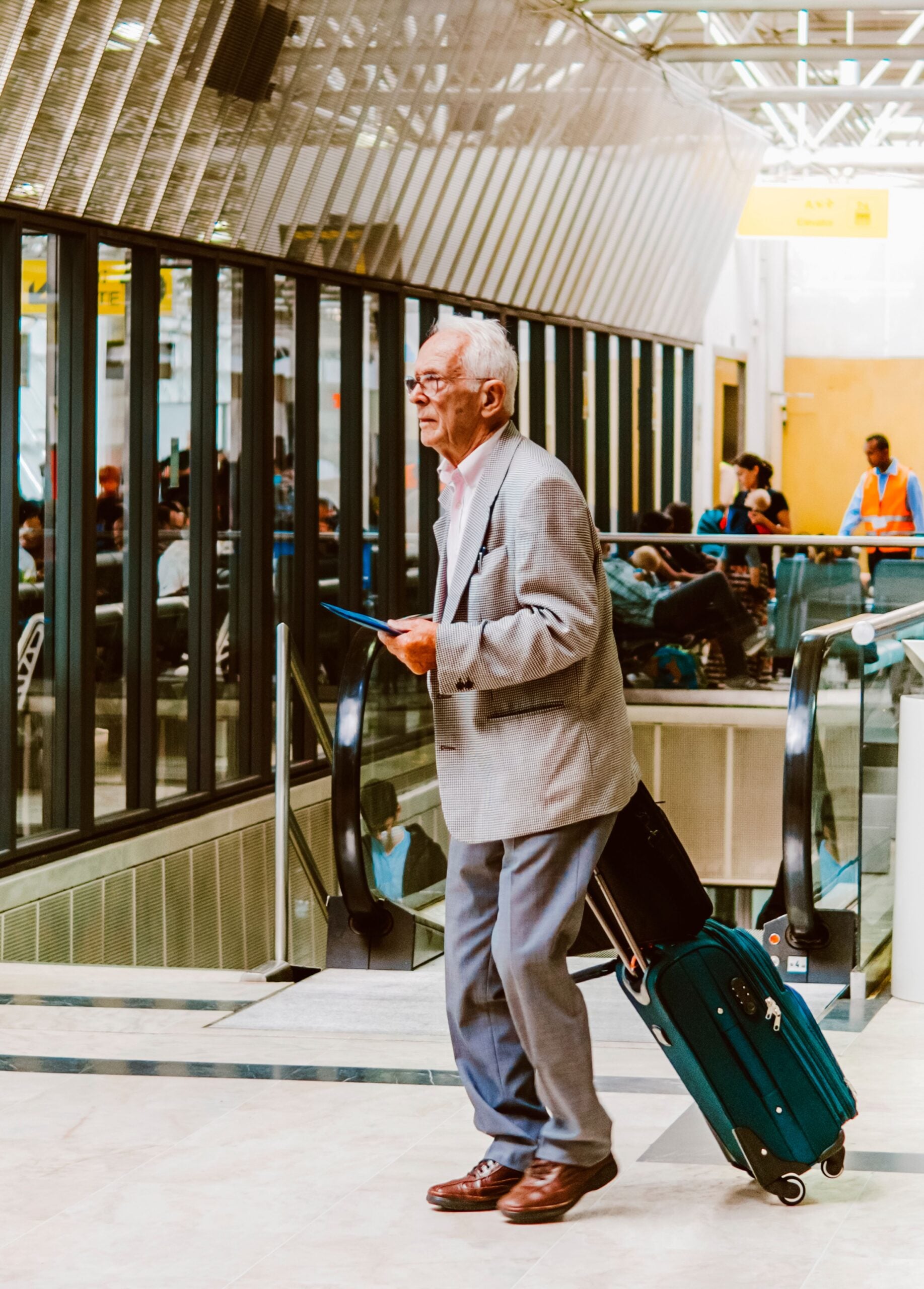 An older man in a gray suit stands in an airport terminal, holding a tablet and pulling a teal suitcase. People sit behind glass windows; an escalator and airport signs are visible.