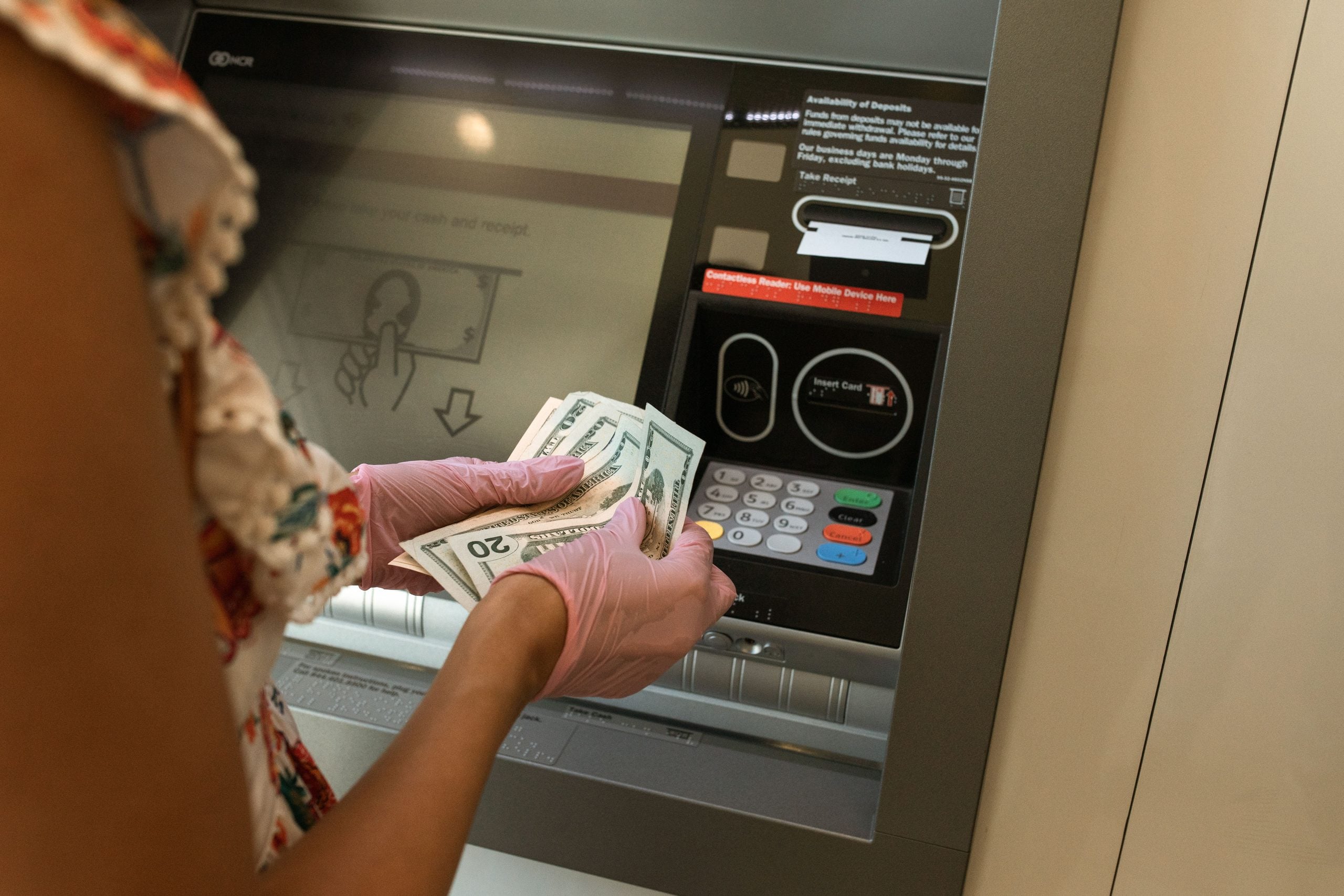 A person wearing pink gloves counts U.S. dollar bills while standing at an ATM. The ATM screen and keypad are visible in the background.