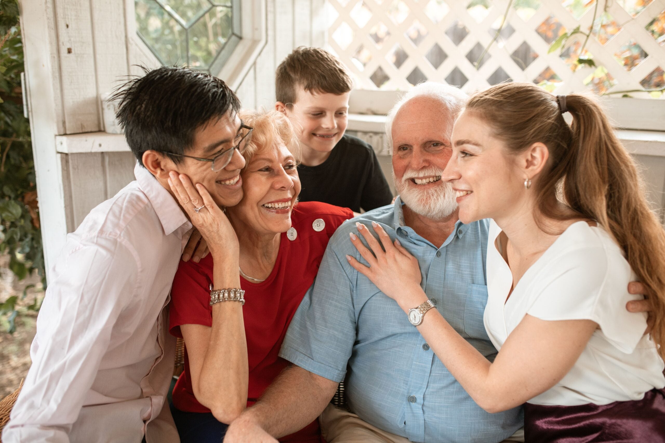 Five people of different ages, smiling and sitting closely together on a porch, appear happy and affectionate, enjoying each other’s company in a bright outdoor setting.
