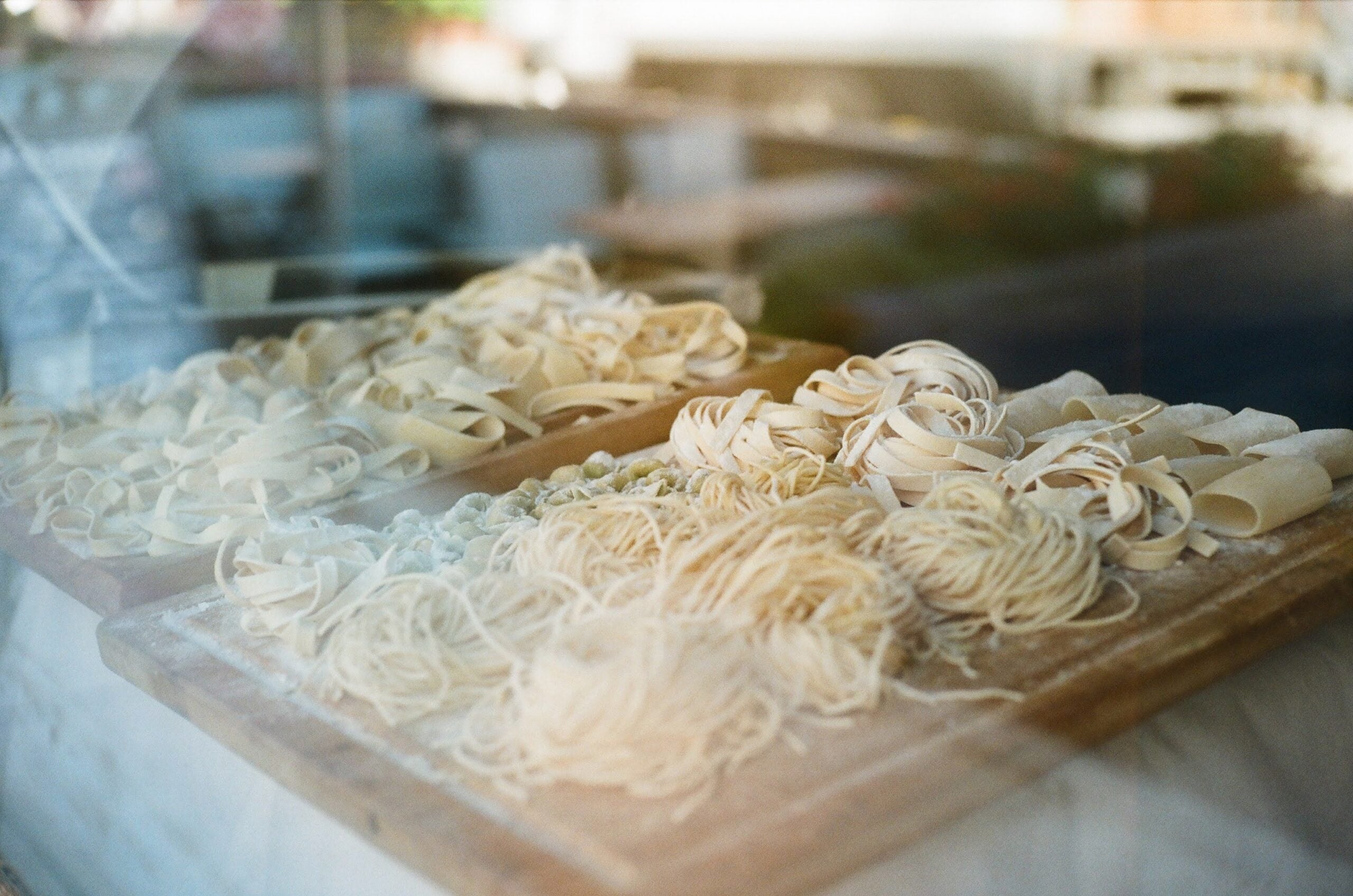 Various types of fresh pasta, including ribbons and nests, displayed on wooden boards behind a window.