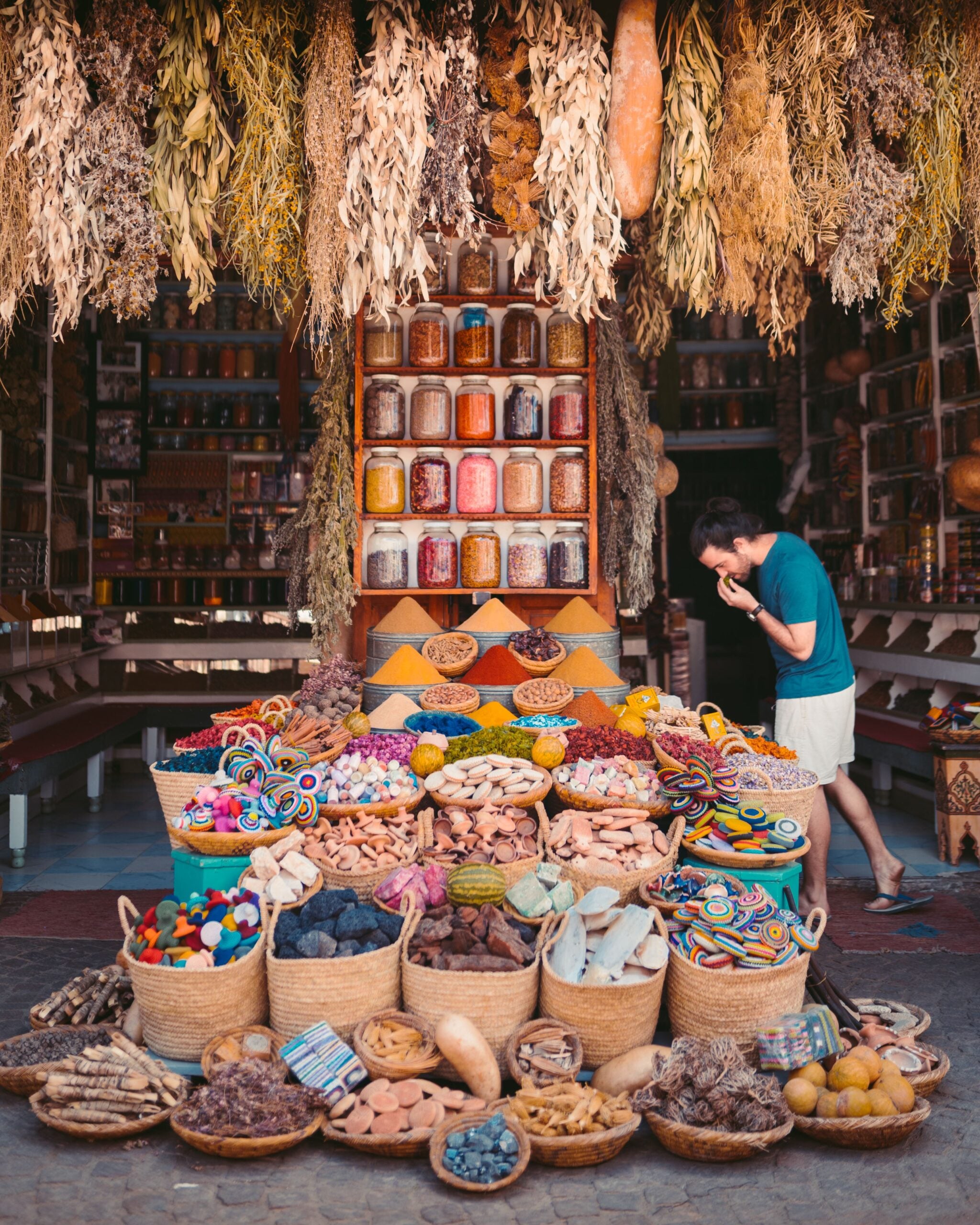 A man smells spices at a shop filled with baskets of colorful powders, dried goods, and rows of glass jars, with bundles of dried herbs hanging from the ceiling.