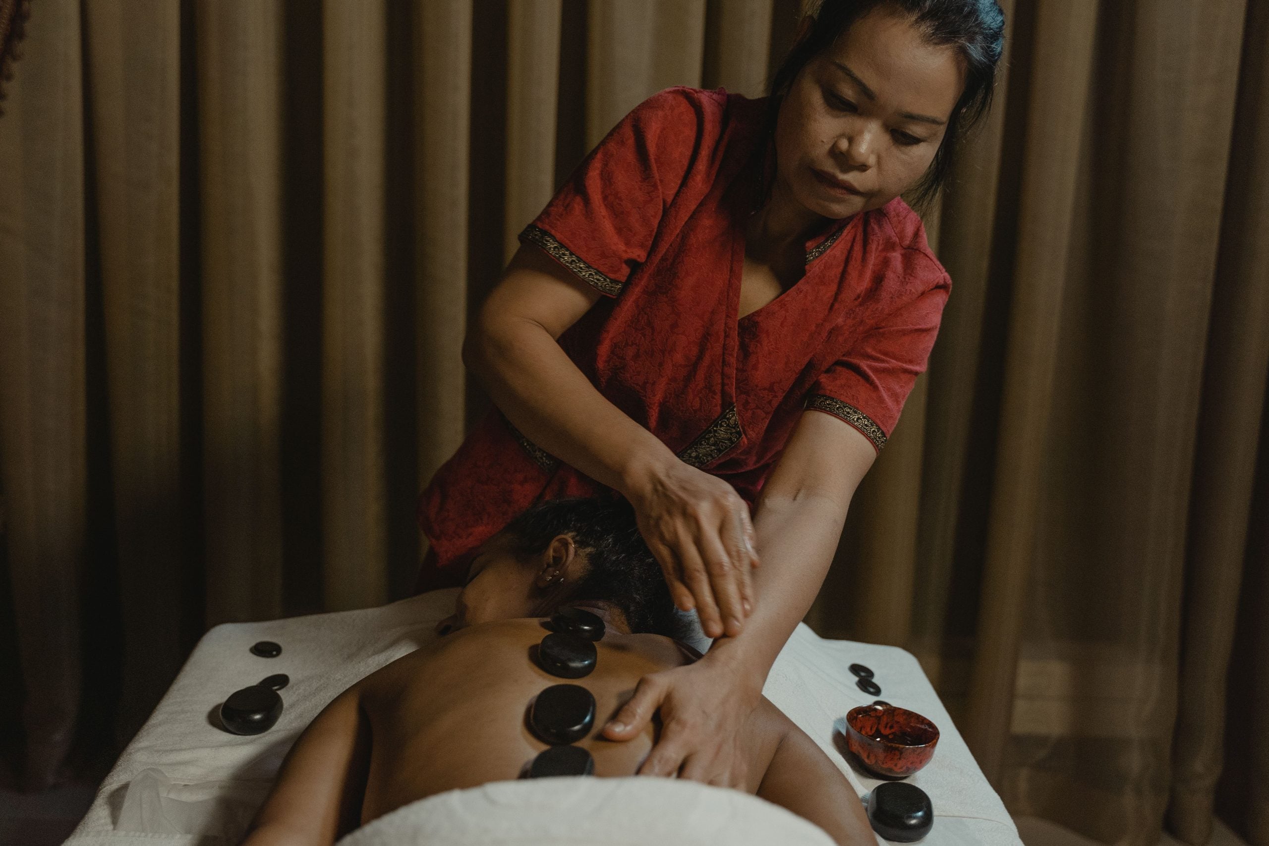 A woman gives a hot stone massage to a person lying face down on a table, with several black stones placed along their back. Brown curtains are in the background.