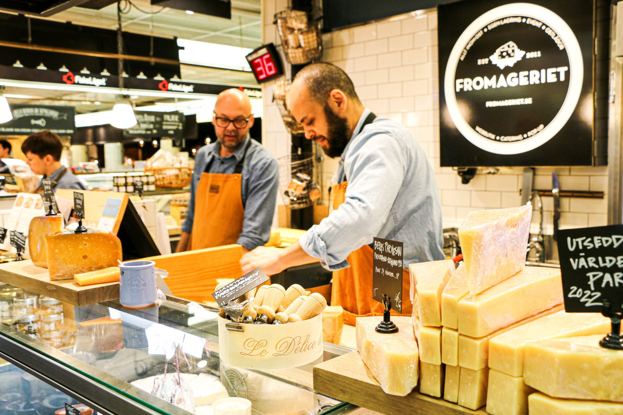 Two cheesemongers wearing aprons work behind a deli counter filled with large blocks and wedges of cheese.