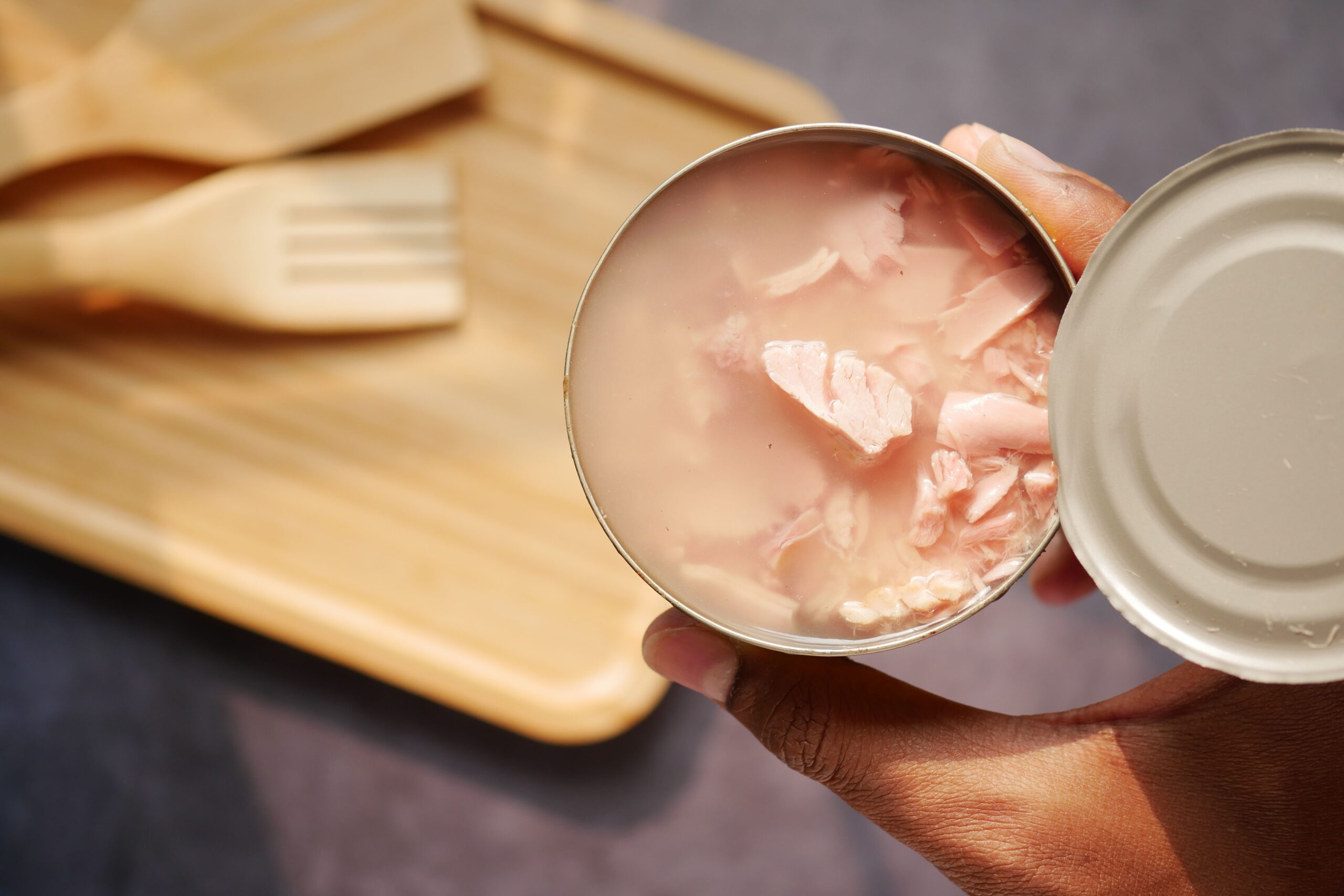 A hand holds an open can of tuna in water above a wooden cutting board and wooden utensils.