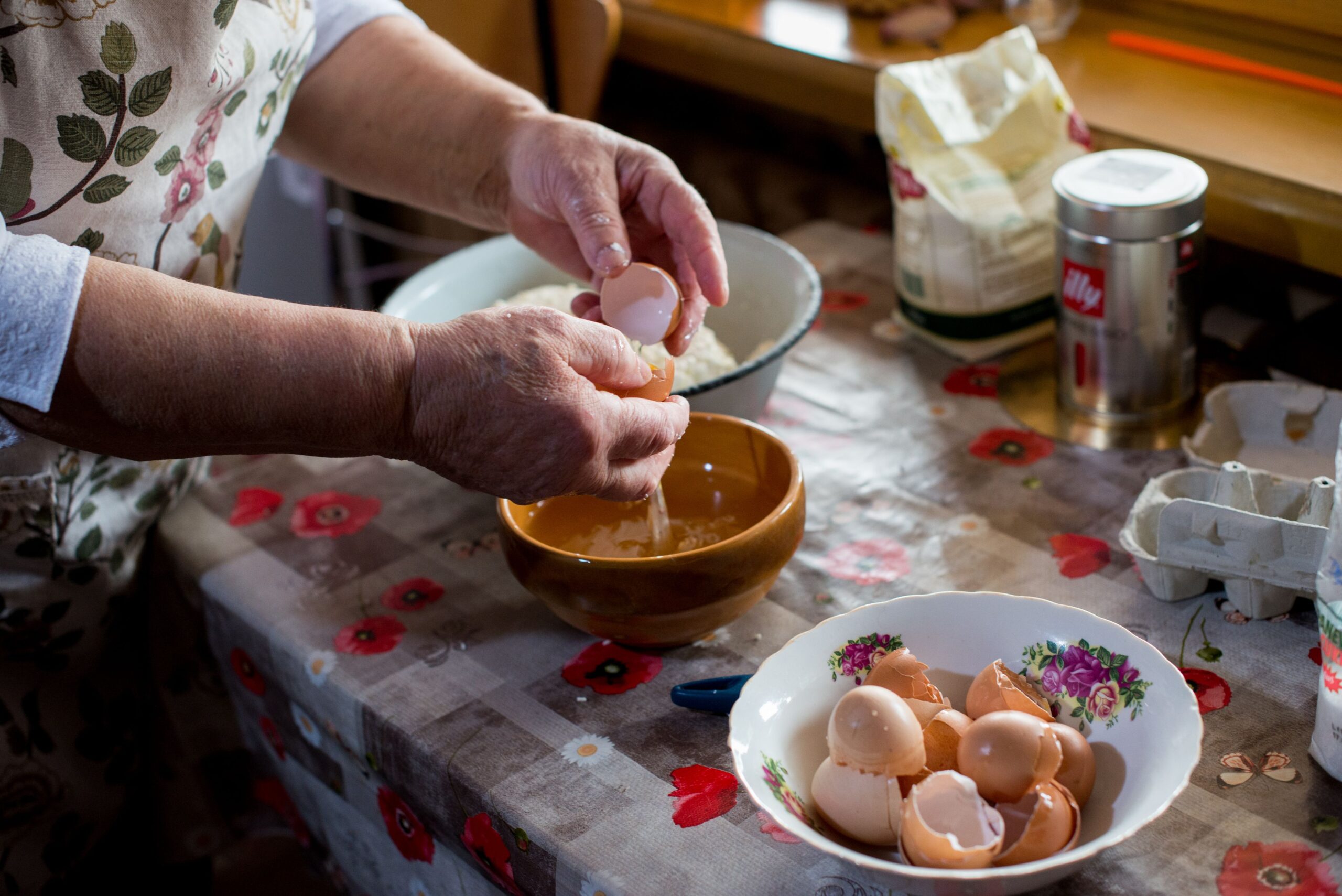 A person cracks an egg over a bowl at a kitchen table covered with ingredients, egg shells, and utensils, preparing to bake.