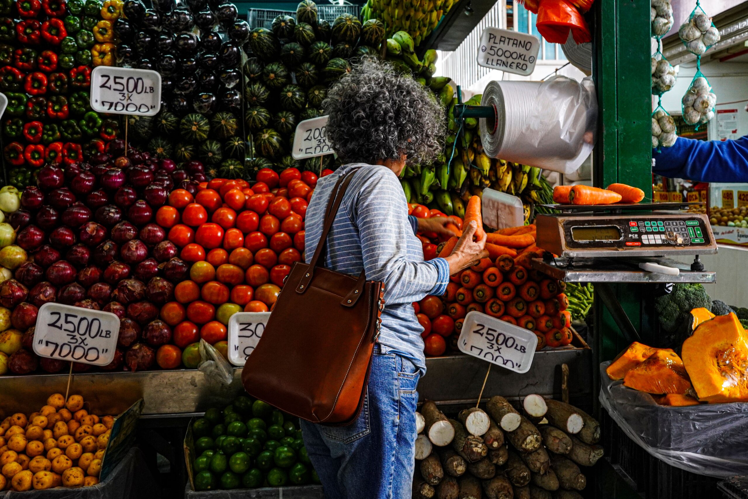 A person with curly gray hair shops at a vibrant fruit stand, holding cash near a weighing scale. Colorful produce like apples, oranges, and bananas is stacked around, with handwritten price signs visible.