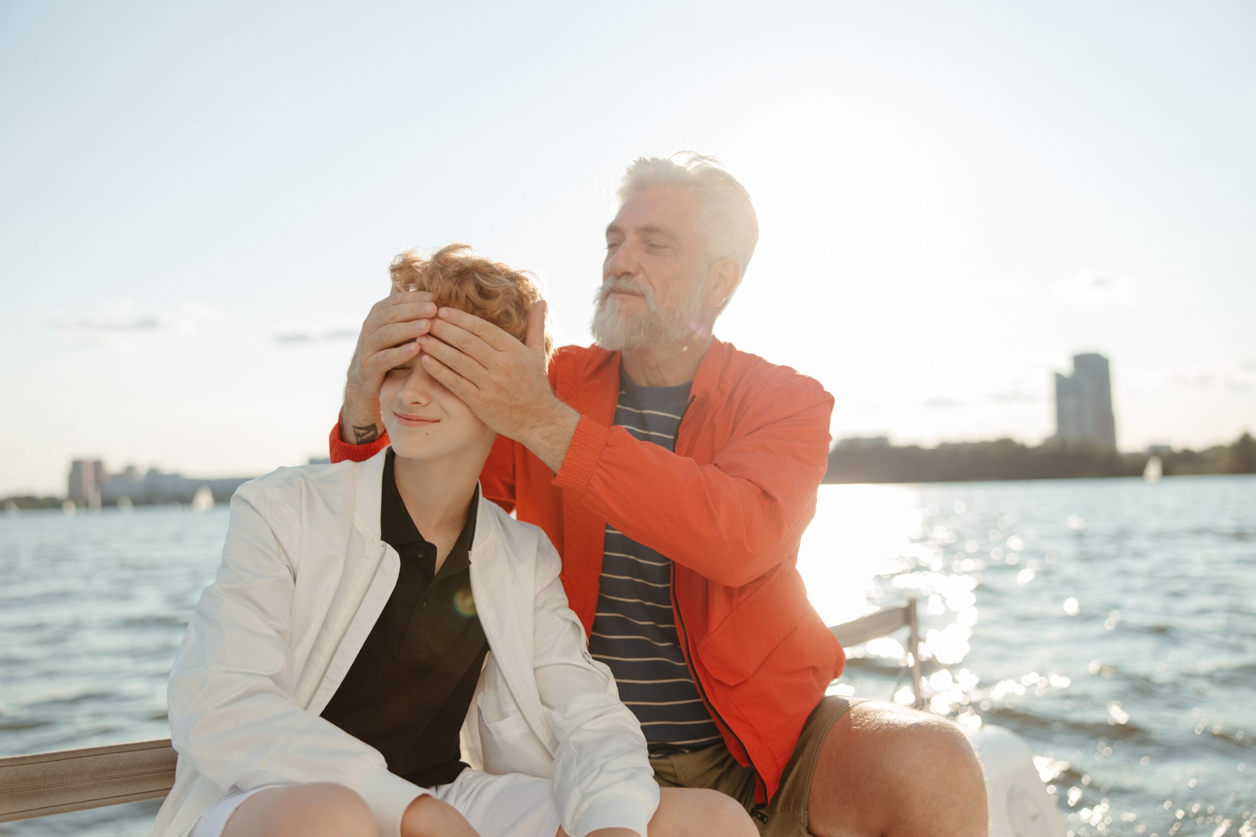 An older man playfully covers a young boy’s eyes while sitting together on a boat, with water and a city skyline visible in the background under bright sunlight.