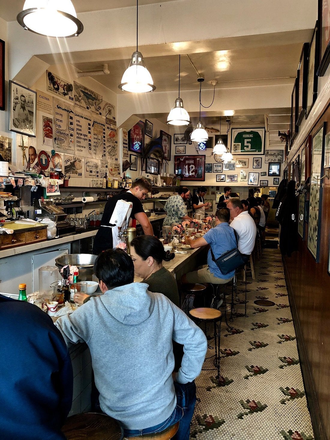 Diners sit at bar seating along a long counter in a narrow restaurant with menus and sports memorabilia displayed on the walls.
