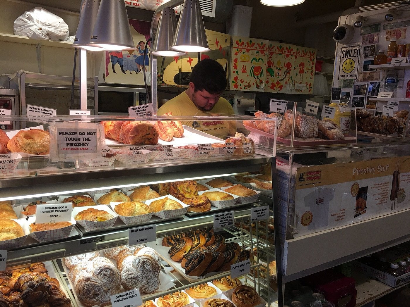 A food display case filled with various piroshky, rolls, and pastries at a bakery counter, with a staff member working in the background.