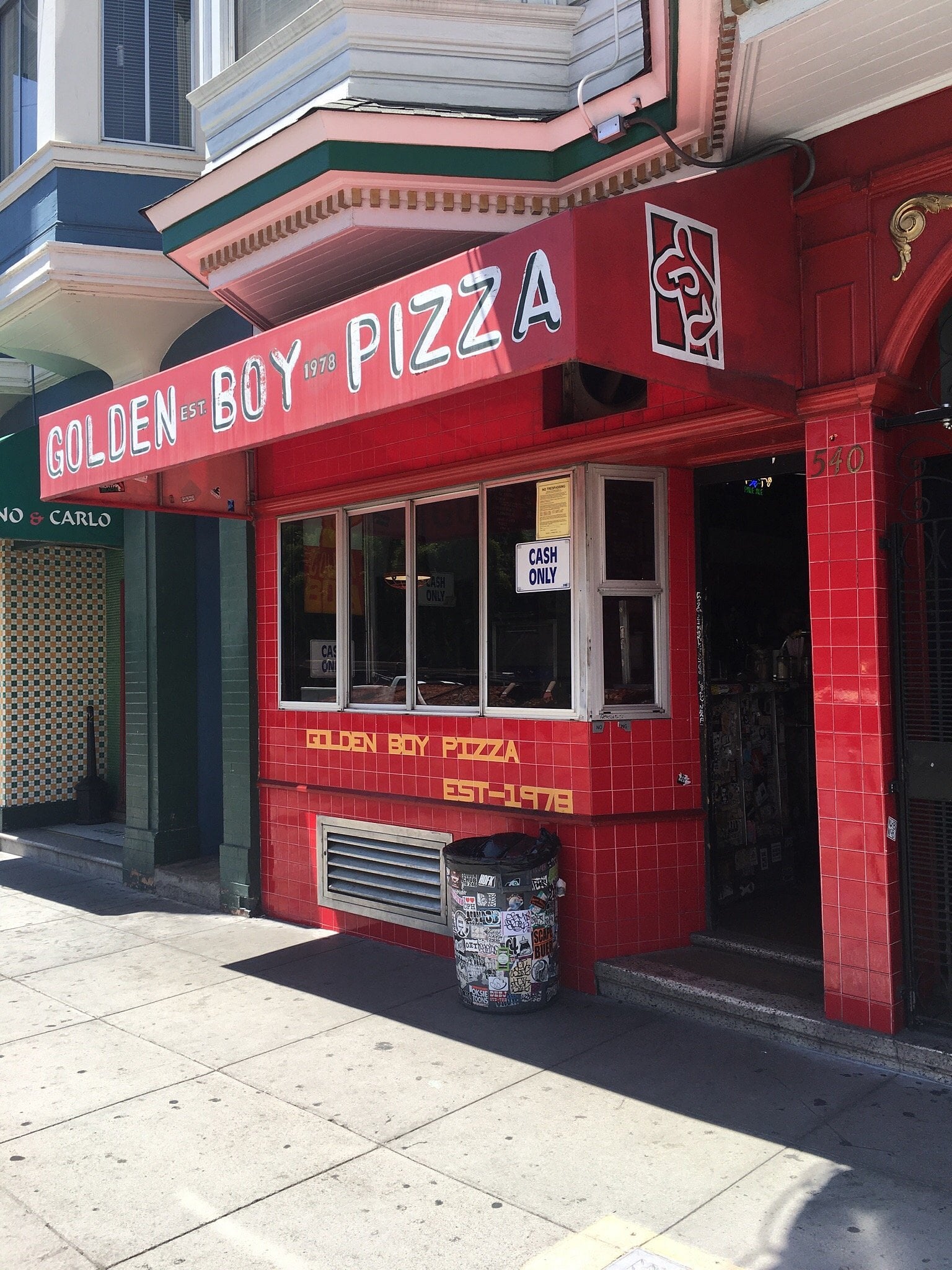 Golden Boy Pizza storefront with a red tiled exterior, a large red awning, and a sticker-covered trash can on the sidewalk.