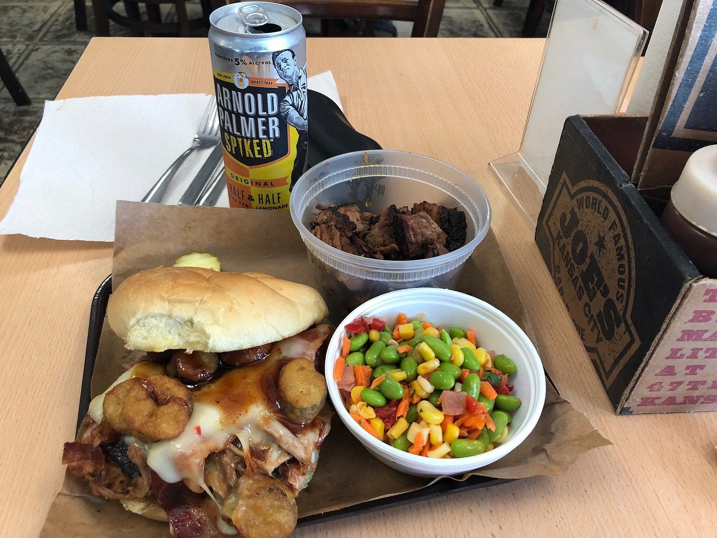 A pulled pork sandwich with pickles and melted cheese, a side of brisket, a bowl of edamame and corn salad, and a can of Arnold Palmer Spiked on a tray.